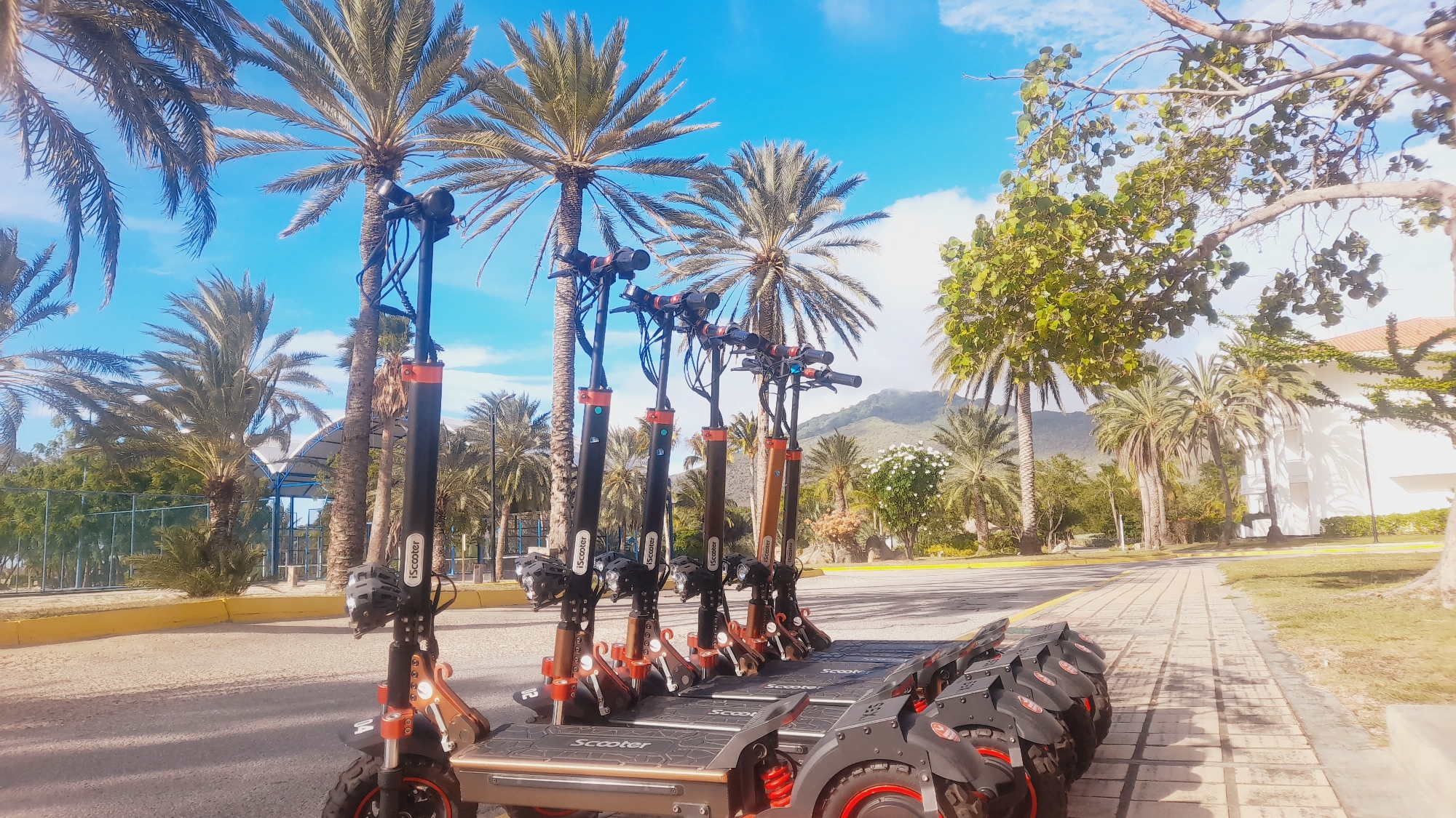 In the city of Pedro González, Venezuela, several electric scooters are parked in a row on a paved walkway. The scooters are predominantly black and orange, with visible branding indicating "iScooter". They are lined up with their handlebars facing upwards. The scene is set against a backdrop of lush tropical vegetation, featuring numerous palm trees of varying sizes, some with their fronds obscuring parts of the bright blue sky, which is dotted with white clouds. In the distance, mountains rise, partially covered by trees. To the right of the scooters, a white building with an orange-tiled roof is visible, bordered by green grass and trees. The ground is a mix of paved walkway and what appears to be a dirt or gravel area near the scooters. The overall impression is a sunny, warm day in a tropical environment.