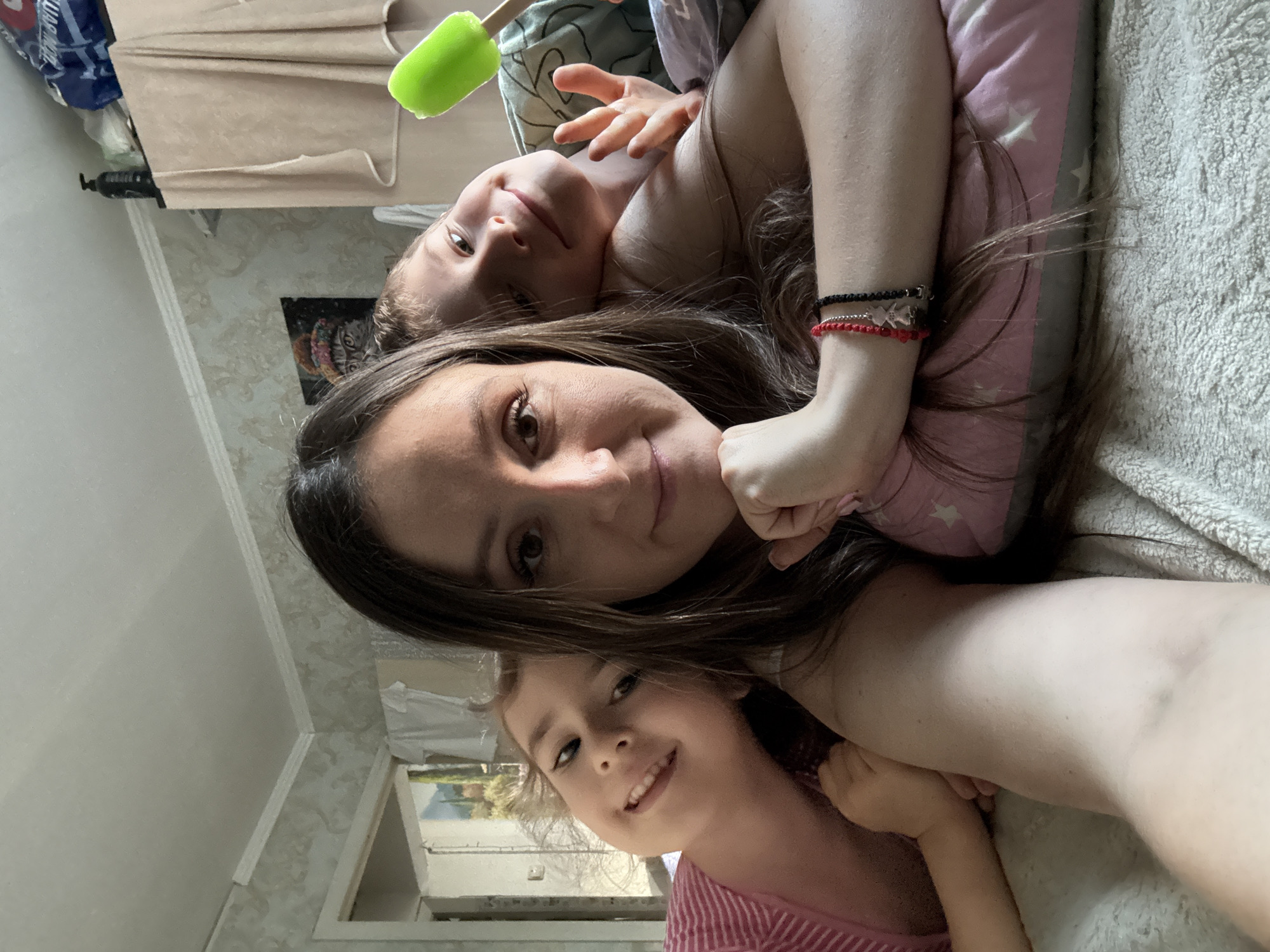 In a domestic indoor setting, a woman and two children are lying on a bed. The woman is in the foreground, taking a selfie with a smartphone. Her left arm is bent and resting on a pink pillow with star patterns. She has long, dark brown hair and is wearing a white top.
To her left, a young girl with blonde hair and a pink striped shirt is leaning her head on the woman's arm. She is looking towards the camera with a wide smile.
To the woman's right, a boy with light brown hair is peeking from behind the woman. He is holding a bright green popsicle in his right hand and has his fingers extended towards the camera. He is smiling and looking at the camera.
The background shows a cluttered bedroom. A doorway on the left reveals a glimpse of another room with a landscape painting. On the right, a wardrobe is visible with towels draped over it, and a black bottle is on top. The room has white molding along the ceiling and patterned wallpaper. The overall impression is a casual, familial moment captured in a typical home environment, likely in Kroyanskoye, Russia, given the context.