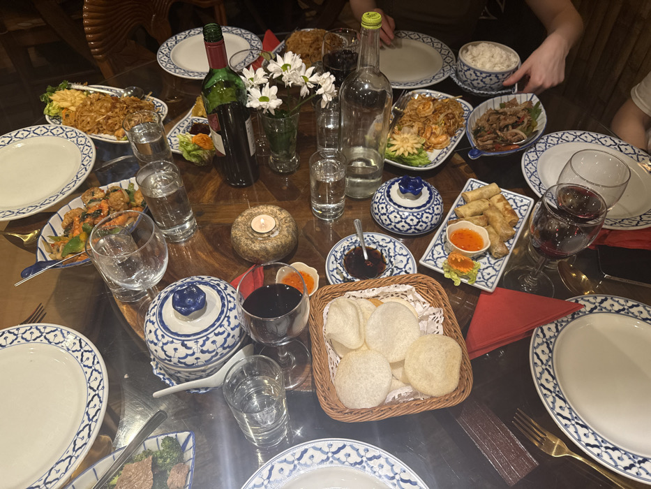 The scene depicts a dinner table setting within a restaurant in Deal, United Kingdom. The dark, polished wooden table is laden with numerous dishes and tableware, suggesting a shared meal for multiple individuals.
**Subjects and Objects:**
The table features an array of white ceramic plates with a distinct blue, intricate pattern, some empty, some in use. Corresponding serving dishes in the same blue and white pattern hold various Thai-style foods. These include:
* A wicker basket containing several light-colored, round prawn crackers.
* A rectangular plate with stir-fried meat and green vegetables (possibly broccoli).
* A plate with noodles (possibly Pad Thai) and shrimp.
* A rectangular plate presenting several crispy spring rolls, accompanied by two small dipping bowls with orange and red sauces.
* A small circular bowl filled with a dark dipping sauce.
* A bowl of white rice.
* A lidded, ornate blue and white ceramic pot, possibly for soup or rice, with a white ceramic spoon resting inside.
* A plate with fried appetizers or small cakes, garnished with greens.
Beverages include two bottles of red wine, one appearing full, the other partially consumed. Multiple clear drinking glasses are present, some holding water, others empty or containing red wine. An empty glass bottle is also on the table.
**Decorations and Other Items:**
A small, lit candle in a textured, light brown holder provides ambient lighting. A slender vase contains a small bouquet of white daisies. Red cloth napkins are folded on the table. Gold-colored forks are visible next to some place settings.
**People and Interactions:**
At least two individuals are partially visible through their hands and forearms. One person's hands are observed on the right side of the image, appearing to hold or move a bowl of rice. Another person's arm is visible towards the bottom right. The arrangement indicates an ongoing or recently concluded communal dining experience.