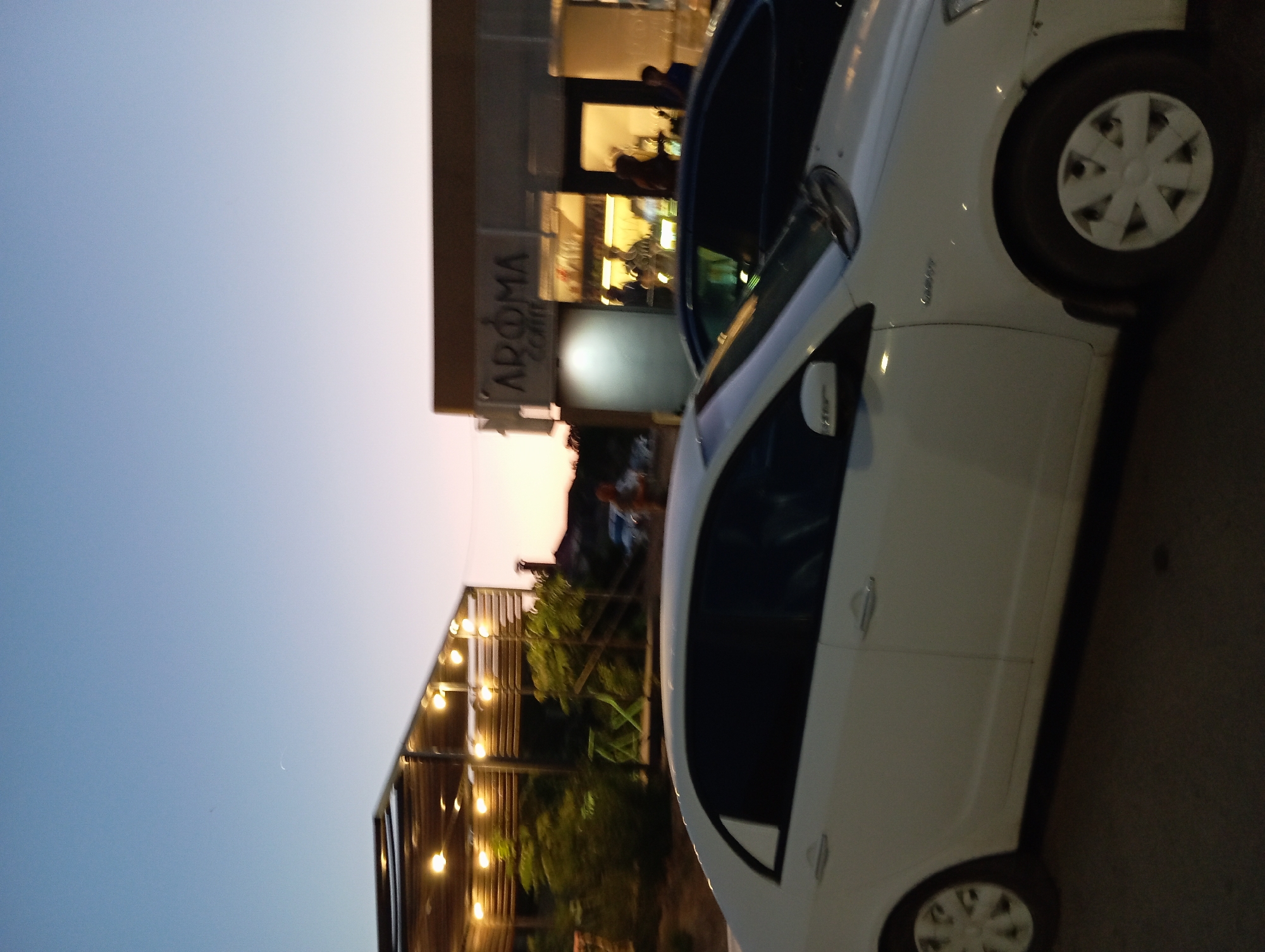 The image shows an outdoor scene at dusk, likely in the city of Aktau, Kazakhstan. In the foreground, a white car is parked. Behind the car, there is a building with a sign that reads "AROMA COFFEE". Several people are visible inside and outside the coffee shop. To the left of the coffee shop, there is an outdoor seating area with lights. The sky is a gradient of blue and orange, indicating the setting sun. The overall impression is a casual evening at a coffee shop in a city.