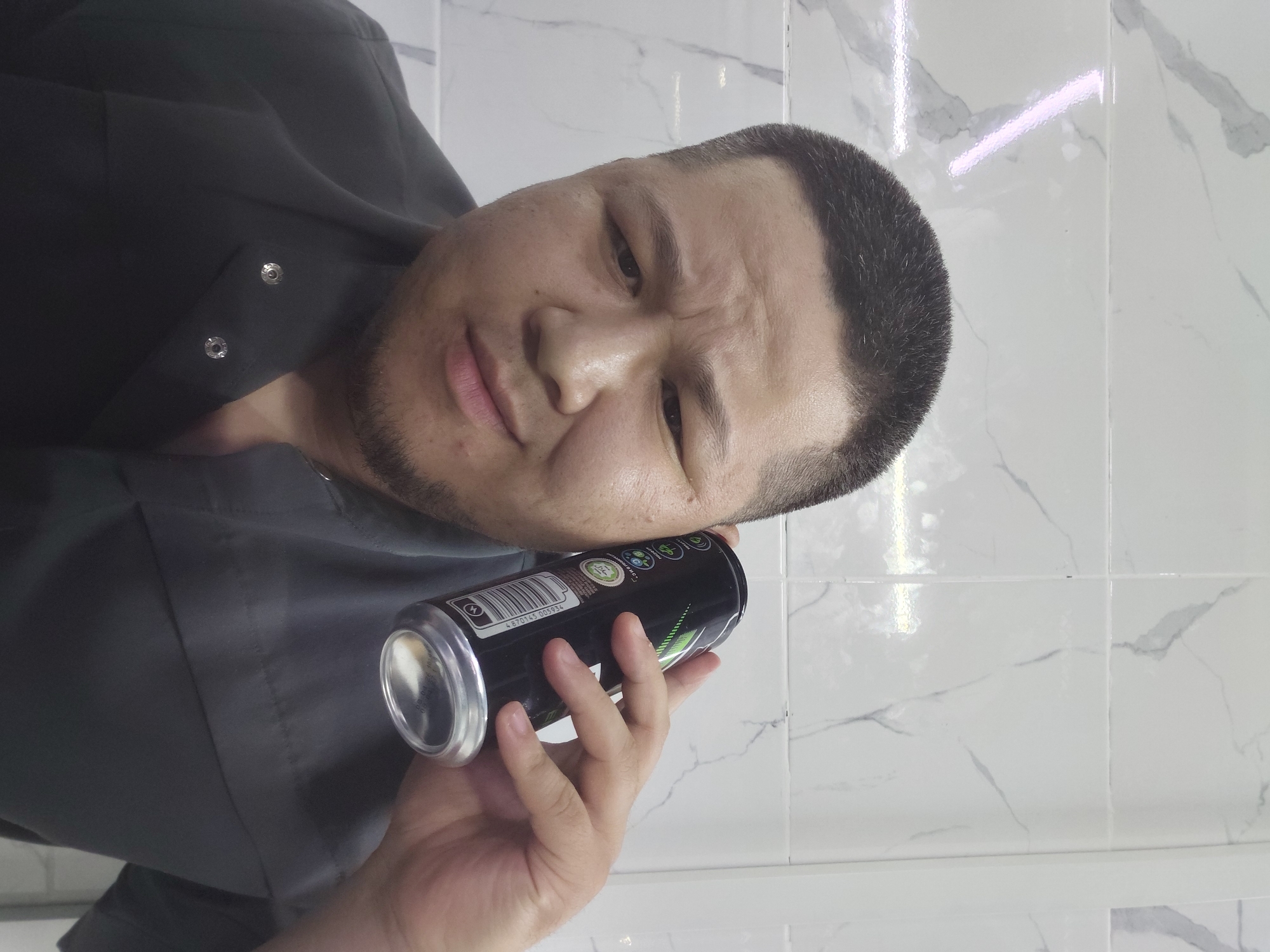 The media file shows a man holding an energy drink can to his ear. He is wearing a dark grey button-up shirt, and the background appears to be a tiled wall, possibly in a bathroom. The man has short dark hair and a beard. His expression is difficult to interpret due to the angle of the photo, but he appears to be concentrating or listening intently. The can is black with green and white text and graphics. Visible text on the can includes a barcode, the number "4870145 005934", and a "Halal" symbol. The lighting suggests it is daytime, and the setting could be indoors. The image is taken from a low angle, looking up at the man.