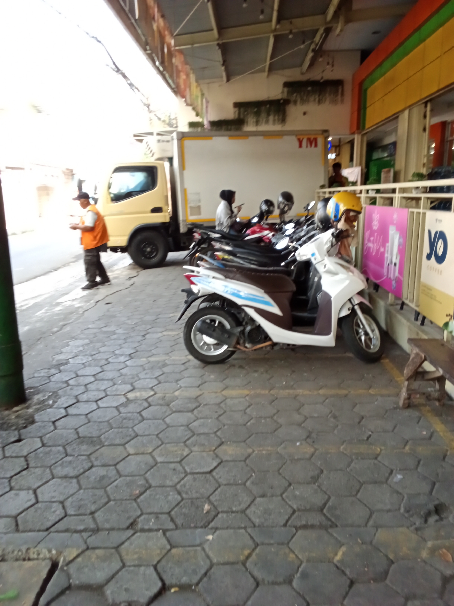 A low-angle, full-shot photograph captures a street scene in Bandung City, Indonesia. In the foreground, a pale yellow truck is parked on the left side of the image, with its rear section facing the camera. To the right of the truck, a white scooter with brown accents is parked parallel to the sidewalk. A yellow helmet rests on the front of the scooter. Behind the scooter, a row of motorcycles is visible, along with a tall, dark green cylindrical object on the far left, possibly a pillar. A man in an orange vest and black pants is walking away from the truck towards the right side of the image. The ground is paved with hexagonal grey bricks. In the background, a building with yellow and orange accents is visible, along with a sign displaying the letters "YO COFFEE". The image is taken during the day, with bright sunlight casting shadows on the ground.