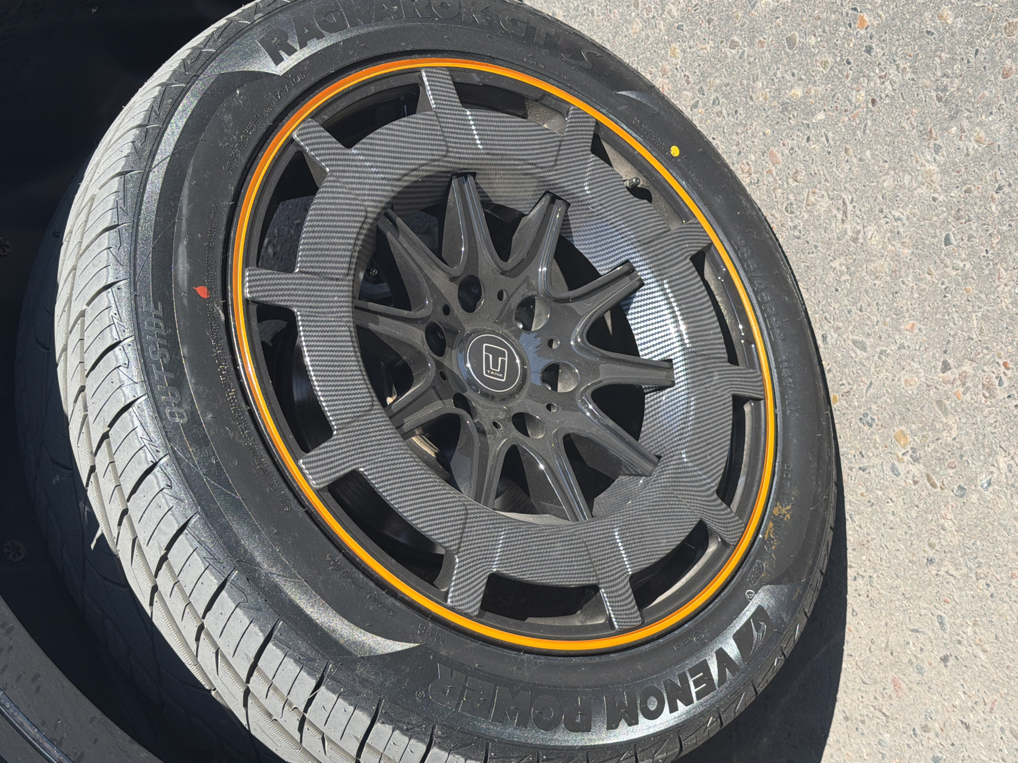A close-up, slightly angled shot shows the side of a car tire and wheel. The tire is black with white lettering, including "VENOM POWER" and "OUTSIDE". The wheel is dark gray with a complex, multi-spoke design. A prominent orange stripe runs along the edge of the wheel. The wheel spokes have a carbon fiber-like pattern overlaid on them. The tire has a tread pattern visible. The ground beneath the tire is a grey, speckled asphalt surface. Shadows fall across the tire and wheel, suggesting outdoor lighting. There are small, colored dots on the tire sidewall, one red and one yellow. The background is dark, likely the car's fender or bodywork. There are no people visible in the image. The context of Krasnoyarsk, Russia, is not visually ascertainable from the image itself.