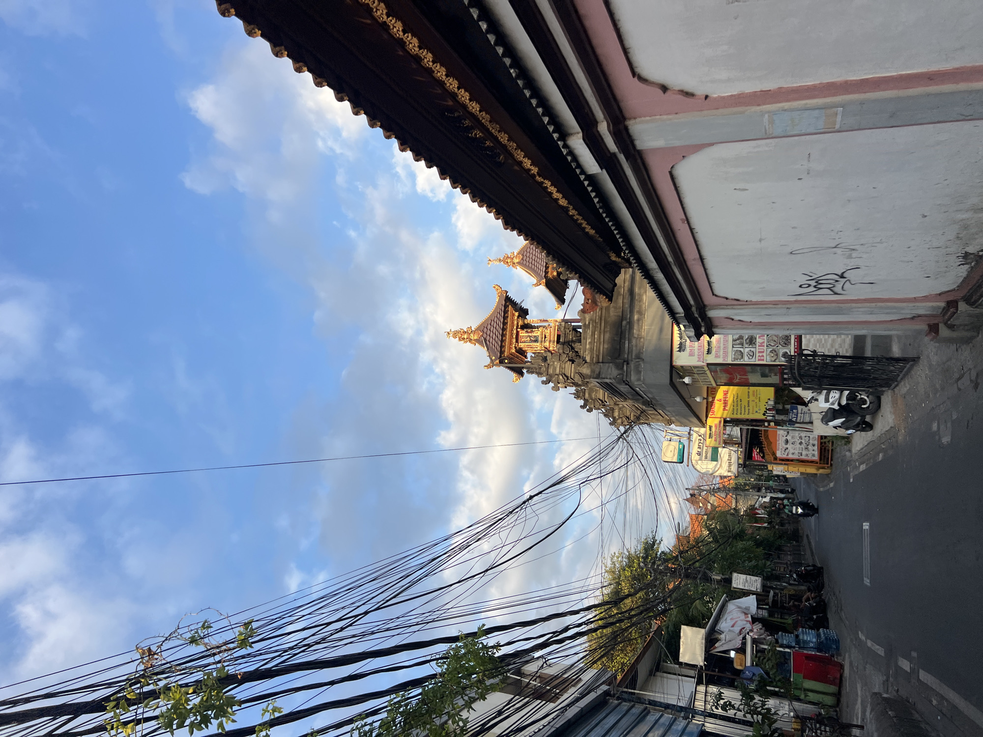 The image shows a street scene in Kuta, Indonesia, with a narrow road bordered by buildings and overhead electrical wires. The sky is partly cloudy with a mix of blue and white. On the right side of the street, a portion of a wall with pink trim and graffiti is visible, with the roof of a building featuring ornate golden detailing extending above it. To the left, buildings with shops and signs line the street, with several parked motorcycles and a few people. Further down the street, more buildings and trees are visible, along with a motorcycle in motion. In the background, traditional Indonesian architectural elements, specifically tiered roofs, can be seen. The overall impression is of a busy urban street with a mix of commercial activity and cultural architecture.