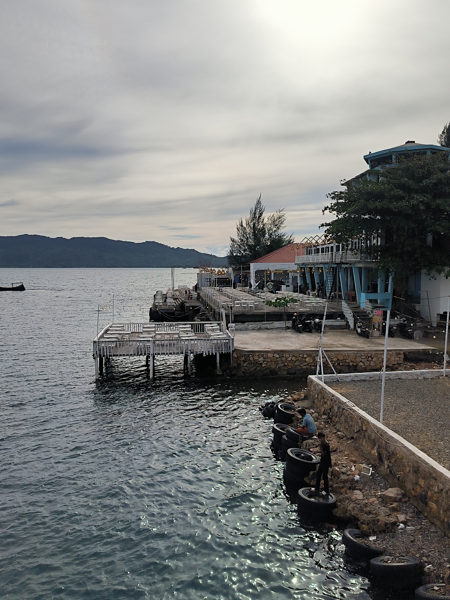This image depicts a waterfront scene, likely a cafe or restaurant, with a view of a lake or sea and distant, hazy mountains. The sky is overcast with soft, diffused light, suggesting either early morning or late afternoon. The water is rippled, reflecting the subdued light.
In the foreground, on a rocky embankment, two young boys are interacting with large black tires. One boy, wearing a blue shirt, is seated on a tire, appearing to be working on something. The other boy, in a black shirt, stands on a tire, looking down. The tires seem to be used as a makeshift barrier or perhaps for some recreational activity.
Behind the embankment, a white wooden pier extends into the water, lined with empty white tables and chairs, suggesting a dining area. Further back, a multi-story building with a light blue exterior and a red-tiled roof is visible, also featuring outdoor seating areas. Several motorcycles are parked on the paved area in front of the building. String lights are visible above some of the seating areas, adding a decorative touch.
The overall impression is one of a relaxed, slightly rustic waterfront establishment. The presence of the people and the setting suggest a typical day in a coastal or lakeside community. There is no visible text in the image.