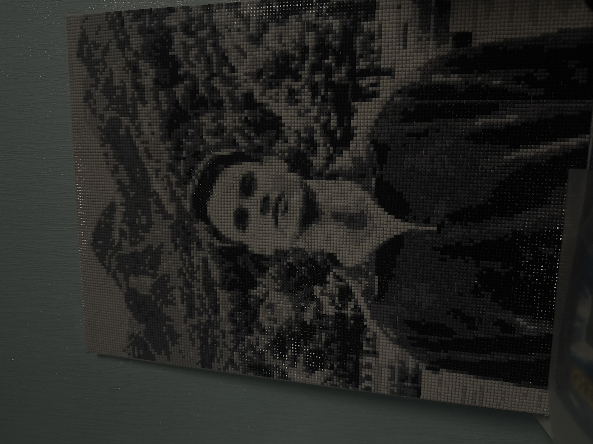 The image shows a pixelated, black and white portrait of a person, possibly female. The portrait is comprised of small, square tiles or pixels, giving it a mosaic-like appearance. The person is wearing a dark outfit, potentially a jacket or coat. The background features a dense, dark texture that may represent foliage or a forest scene. The artwork or item is affixed to a wall painted in a muted green tone. The location, Izhevsk, Russia, cannot be determined from the image.