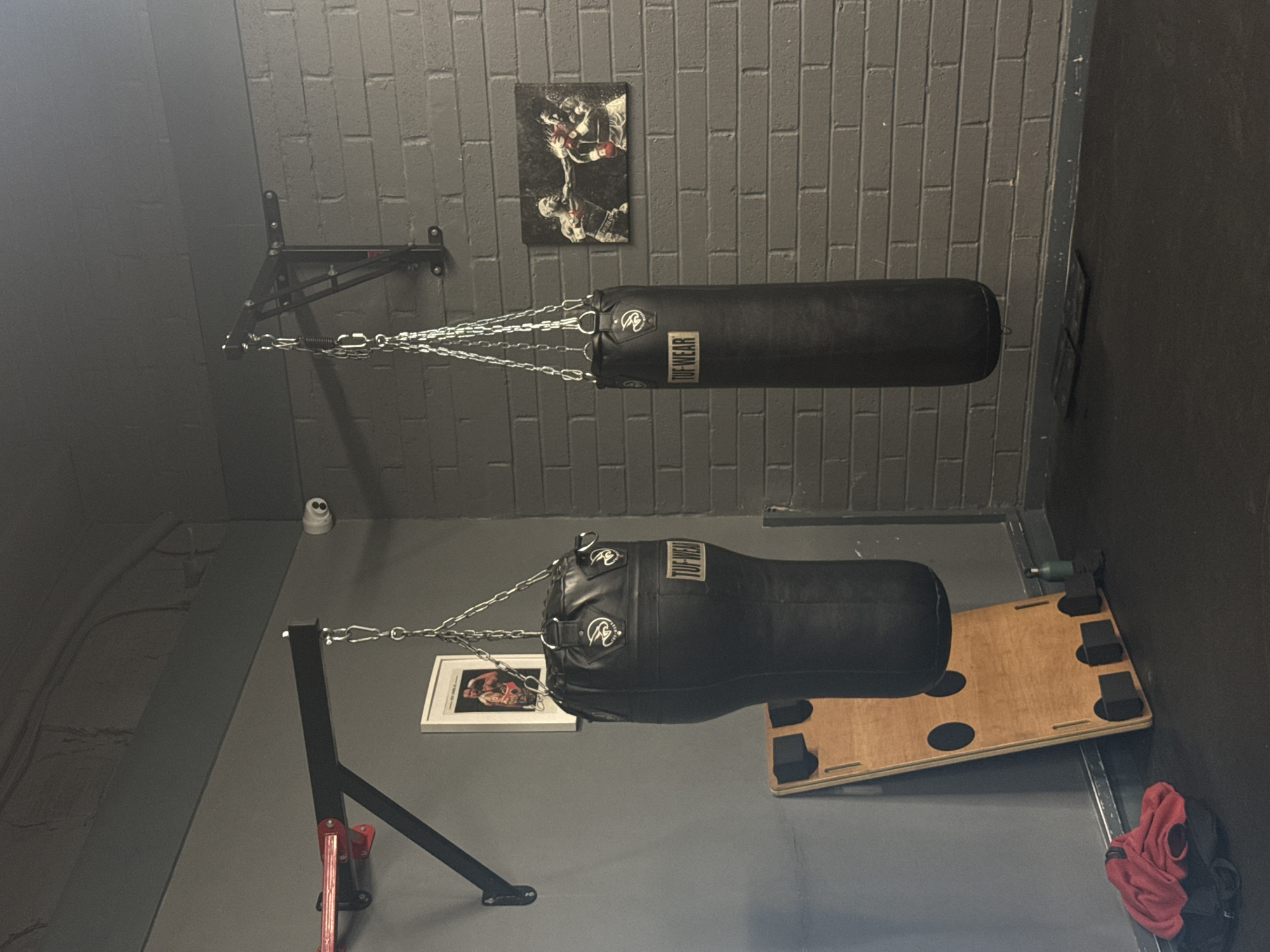 The image shows a gym setting, specifically a boxing or martial arts training area. There are two punching bags suspended from chains, one horizontal and the other vertical. The horizontal bag is suspended from a wall-mounted bracket, while the vertical bag is attached to a floor-standing frame. A piece of wooden equipment, possibly a plyometric box or platform, sits on the floor nearby. A framed photograph or poster rests on the floor near the vertical punching bag. The walls are painted in dark tones, and one wall features exposed brickwork. A small security camera is visible mounted on the wall. A red boxing glove sits in a corner near the wall. The specific location within London, United Kingdom, cannot be determined from the image alone.
