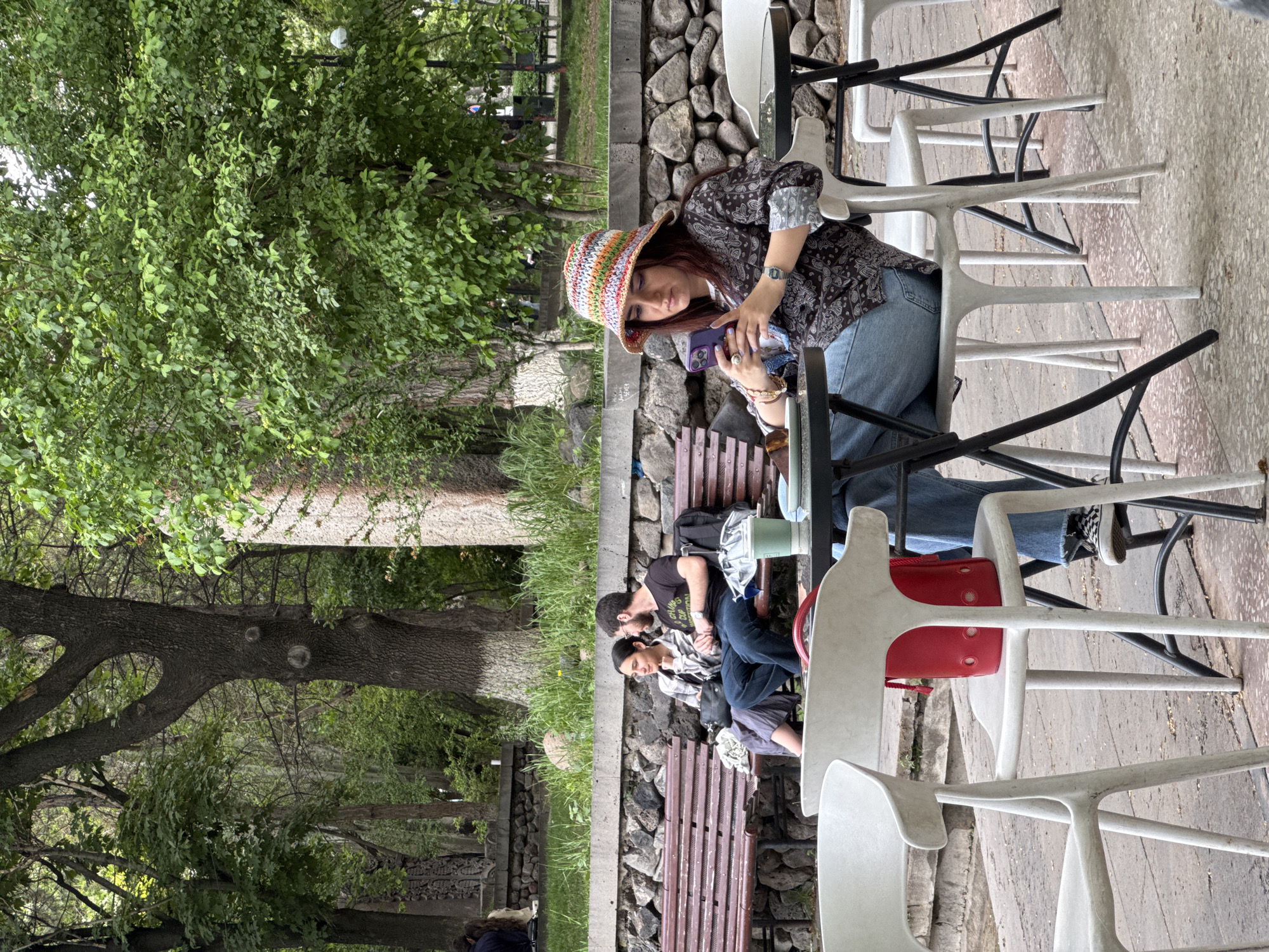 The image depicts an outdoor scene, likely in Yerevan, Armenia, based on the environment and style. A woman in a colorful, patterned bucket hat, a patterned shirt, and jeans is seated at an outdoor table. She is looking at a smartphone in her hands. Adjacent to the table and woman is a bench where a man and woman are seated close together, potentially a couple. The man has a beard, and the woman's head is resting against him. To their left are more people sitting on the same bench. The setting seems to be a park or outdoor café. There are trees and stone walls in the background. Several white and black chairs are scattered around the tables, suggesting an outdoor seating area.