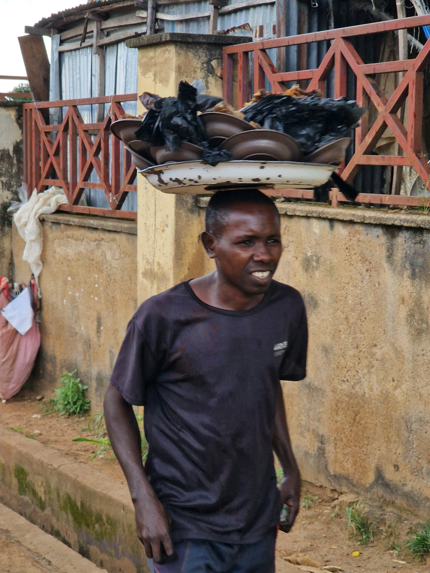 A man is walking outdoors with a tray of items balanced on his head. He is wearing a dark t-shirt and dark pants. The tray is white and has multiple plates stacked on it, covered with black material. The man is looking to his right, and his mouth is open as if he is speaking or yelling. In the background, there is a wall, a fence, and a building with a corrugated metal roof. There is also some vegetation on the ground. The scene appears to be in a developing country, possibly in Africa.