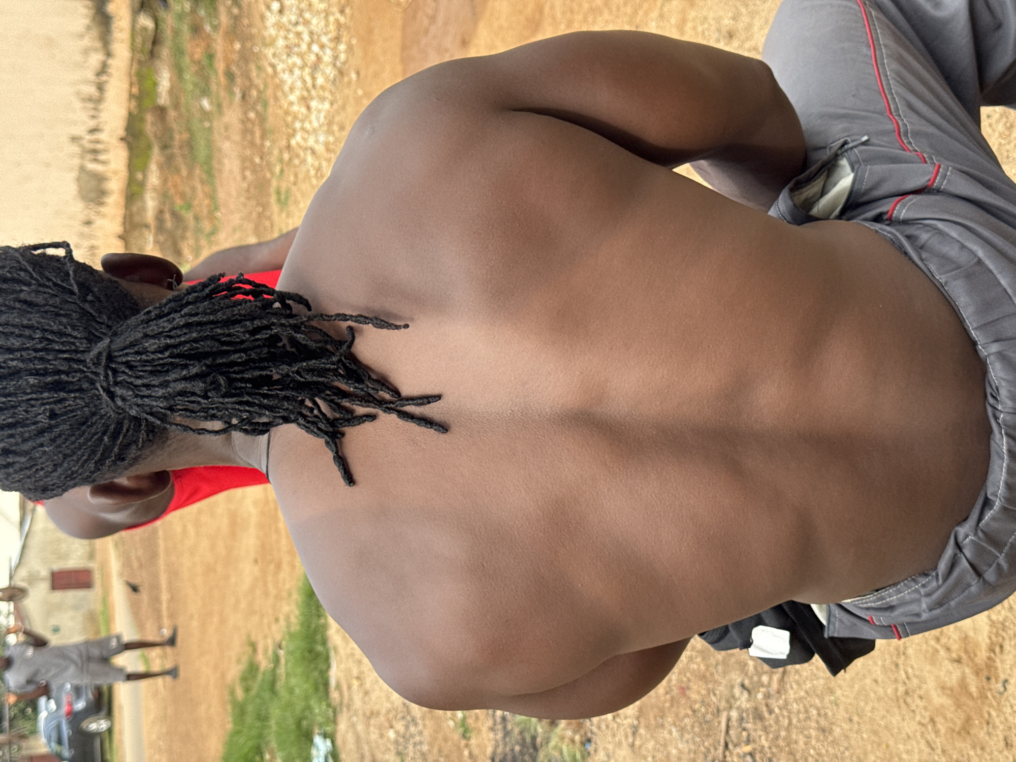 The image, taken in the city of Akwanga, Nigeria, shows the back of a shirtless man with dark, braided hair tied in a ponytail. He is wearing grey pants with red trim. His musculature is visible, particularly his back and shoulders. In the background, another person is visible in the distance, wearing a grey t-shirt and shorts and lifting a weight. The setting appears to be an outdoor area with dirt ground and some grassy patches, with buildings and a car visible further in the background.