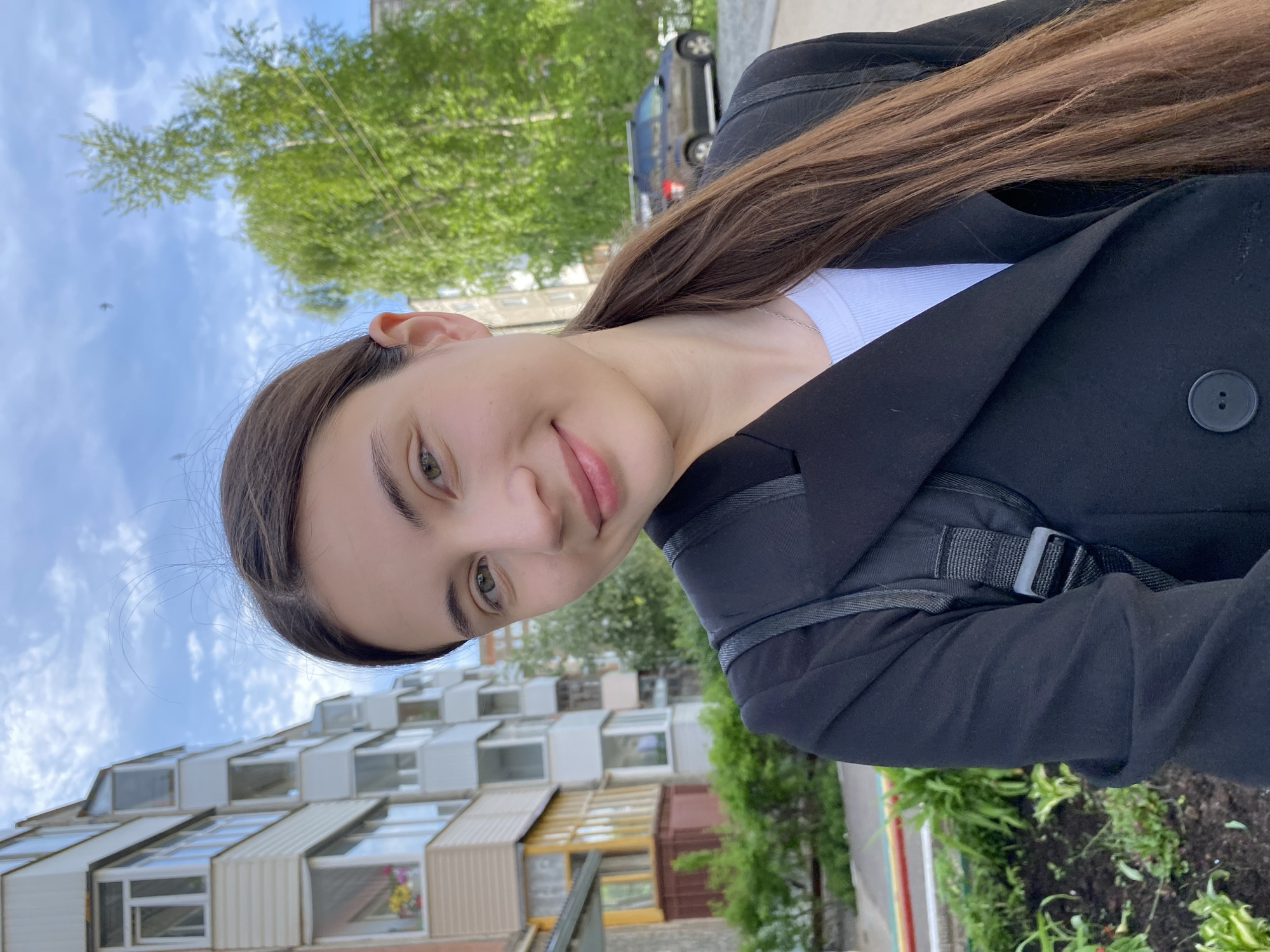 A selfie of a young woman with long brown hair is taken outdoors during daylight hours. She is wearing a black blazer and a white top, and a black backpack strap is visible over her right shoulder. The background features a building with a unique architectural design, possibly residential or commercial. Green trees and a blue sky with some clouds are also in the background. The location is potentially the city of Votkinsk, Russia, given the instructions.