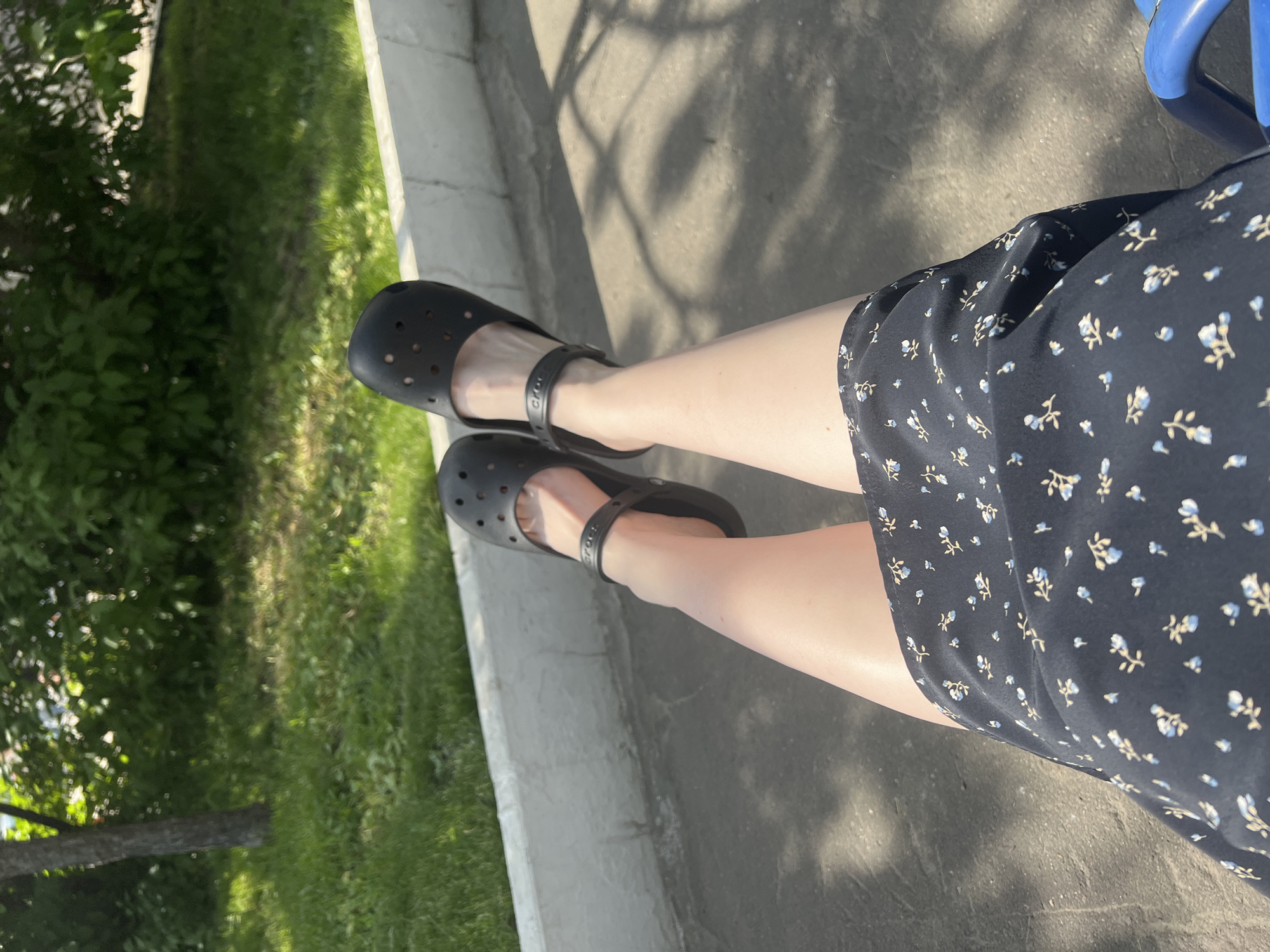 A top-down, medium shot depicts a person seated outdoors. The person is wearing a dark blue dress with small, light blue floral patterns. The person's legs are visible, and they are wearing black Crocs. The background includes a concrete surface with dappled sunlight and shadows, and a strip of green grass and vegetation is visible to the left. The location is in the city of Khimki, Russia.