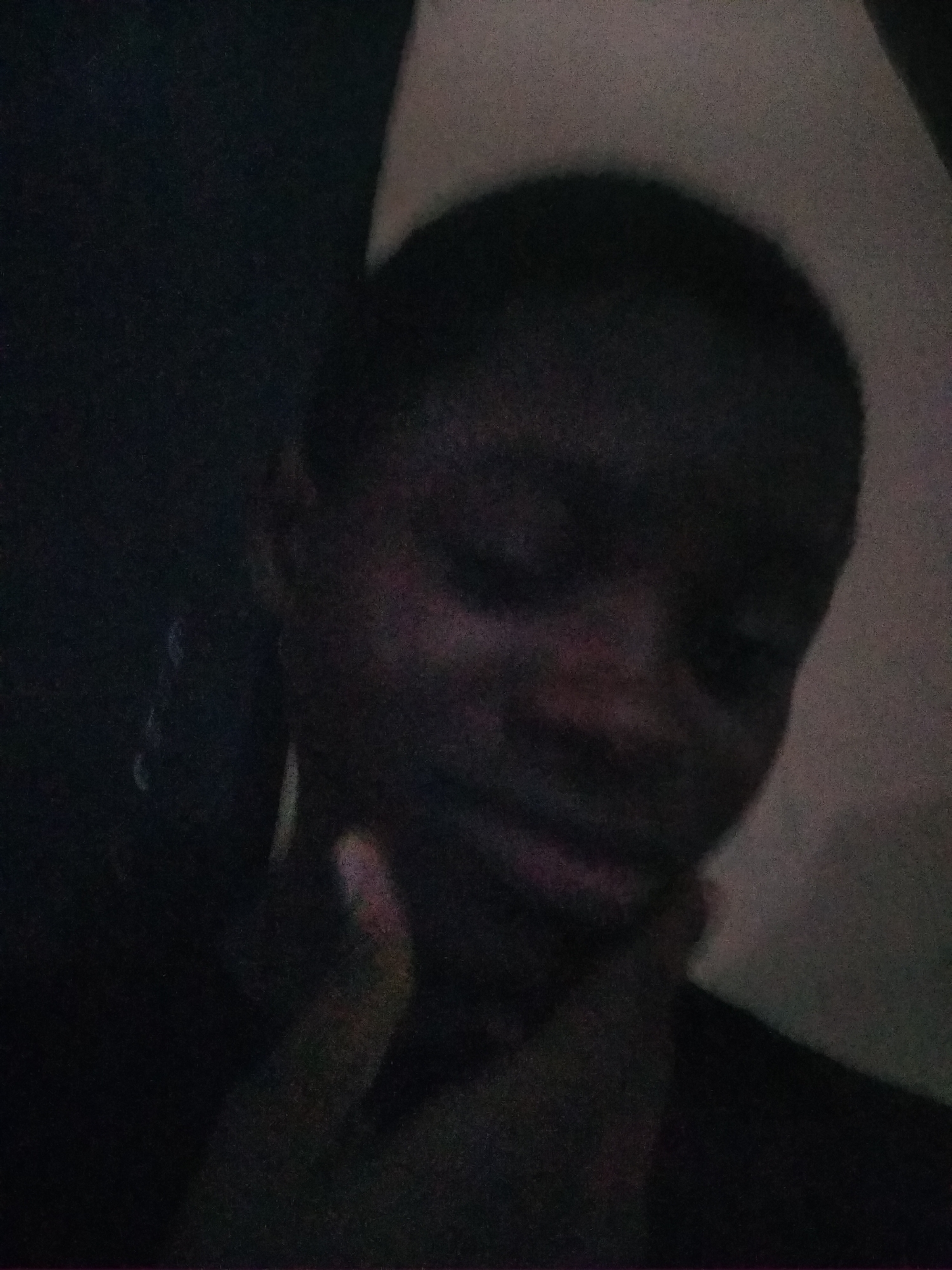 The image shows a person's face, indistinct due to low light and blurriness. A hand is visible touching the face. The background is dark with varying shades of gray, showing an undefined, possibly interior space. Location is unknown.