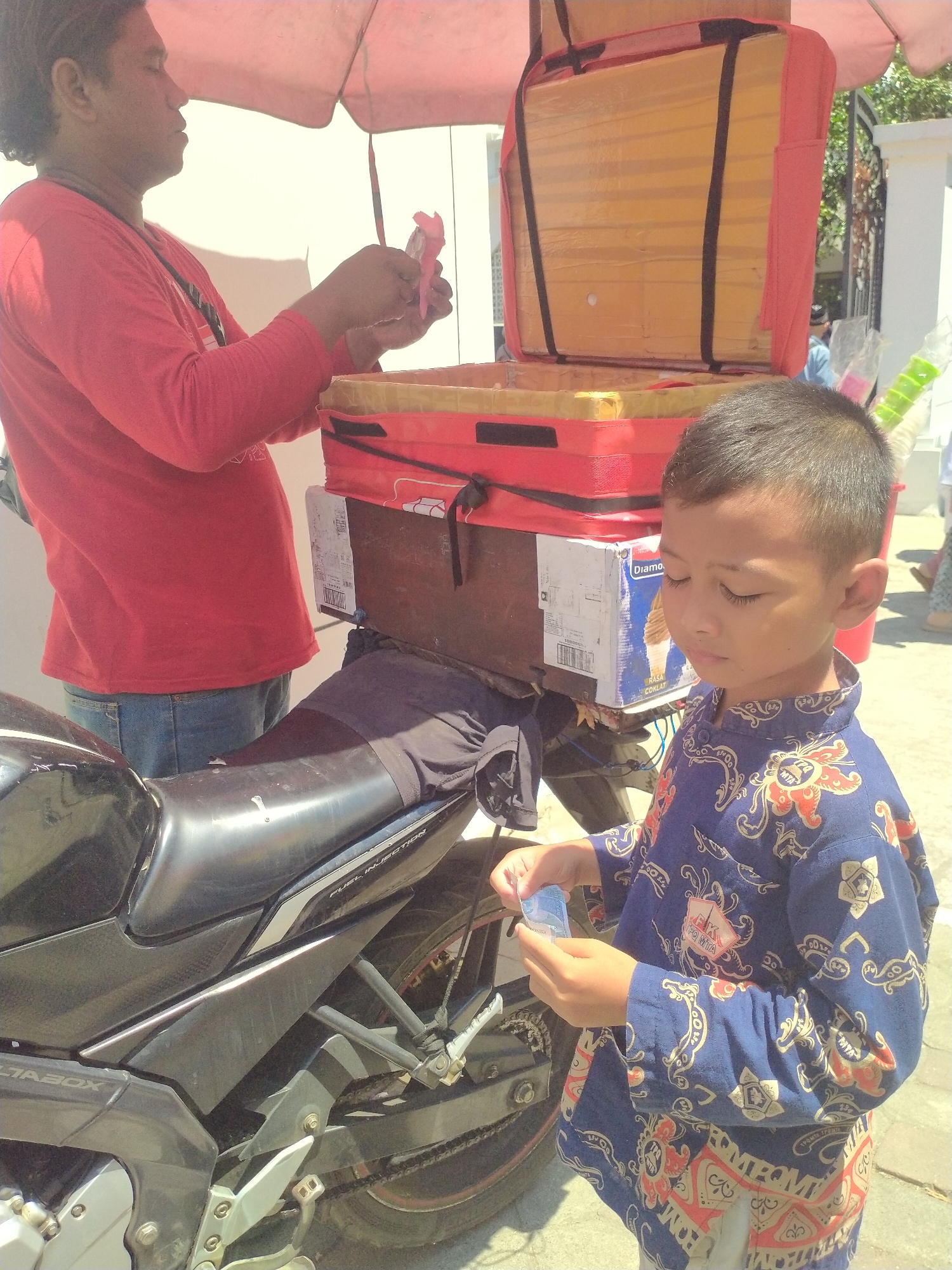 A young Indonesian boy, wearing a blue, patterned shirt, stands next to a motorcycle in Sukoharjo, Indonesia. He is holding a small banknote in his right hand. Behind him, a man in a red long-sleeved shirt stands near a food cart on a bright, sunny day. The cart has a red cooler box on top, and a yellow insulated interior is visible. The motorcycle is black and appears to be a delivery vehicle, with a seat covered by a dark fabric. The background shows a white wall, a metal fence, and some foliage, suggesting an outdoor urban setting. The overall scene depicts a transaction or interaction related to food sales.