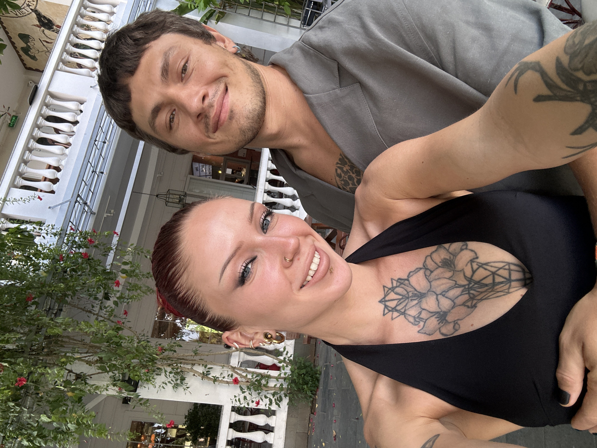 In Hoi An, Vietnam, a couple takes a selfie on a balcony adorned with blooming vines. The woman, with auburn hair, wears a black halter top and shows off a large floral tattoo on her chest, multiple earrings, and nose jewelry. The man, sporting short brown hair and stubble, is dressed in a gray jacket and also has tattoos, visible on his chest and arm. They are smiling and positioned close together, suggesting a romantic encounter. The background features a white railing and more greenery, typical of the charming architecture found in the ancient town.