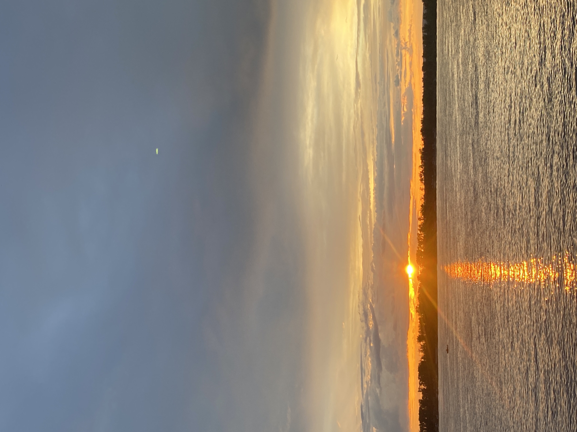 The image depicts a sunset over a wide body of water, likely a river, with a distant tree line on the horizon. The sky is a gradient of colors, with dark clouds at the top transitioning to lighter shades of orange and yellow near the horizon where the sun is partially obscured. A bright reflection of the sun shimmers on the water's surface. In the distance, a small boat can be seen on the water. The environment suggests a natural setting, possibly on the outskirts of a city or town. The specific location in Novokuybyshevsk, Russia, cannot be determined from the image alone.