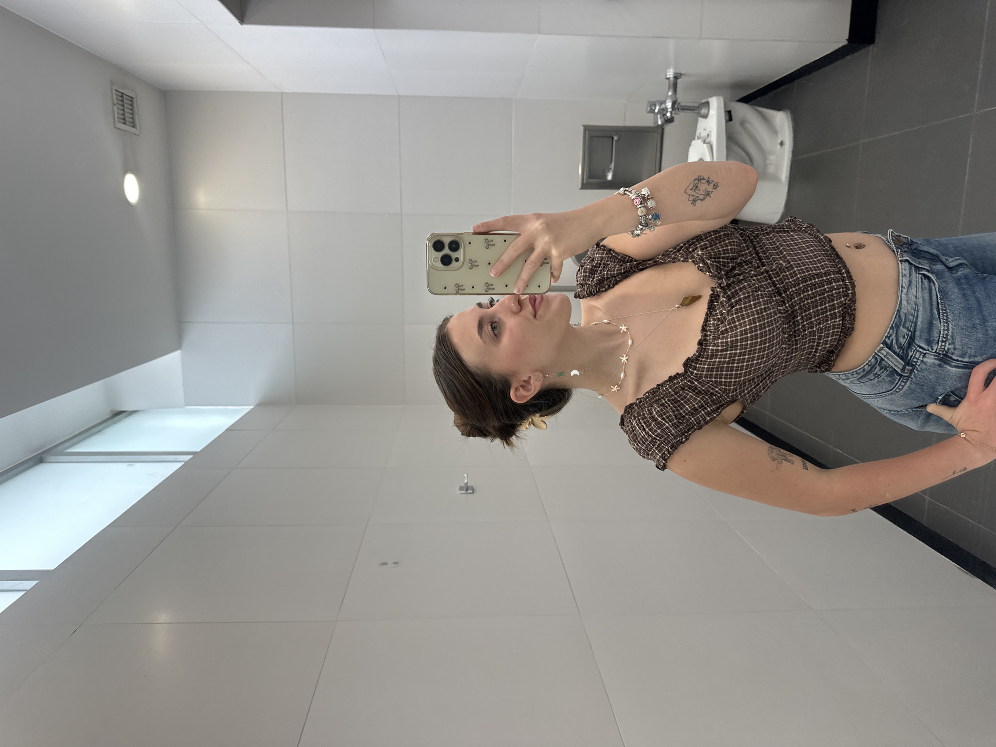 A woman is taking a selfie in a bathroom with white tiled walls and dark gray tiled floors. She is wearing a brown and white checkered crop top, blue jeans, and jewelry. The bathroom has a toilet, sink, and paper towel dispenser. The location is likely a public bathroom, potentially in Mexico City, Mexico, given the request.