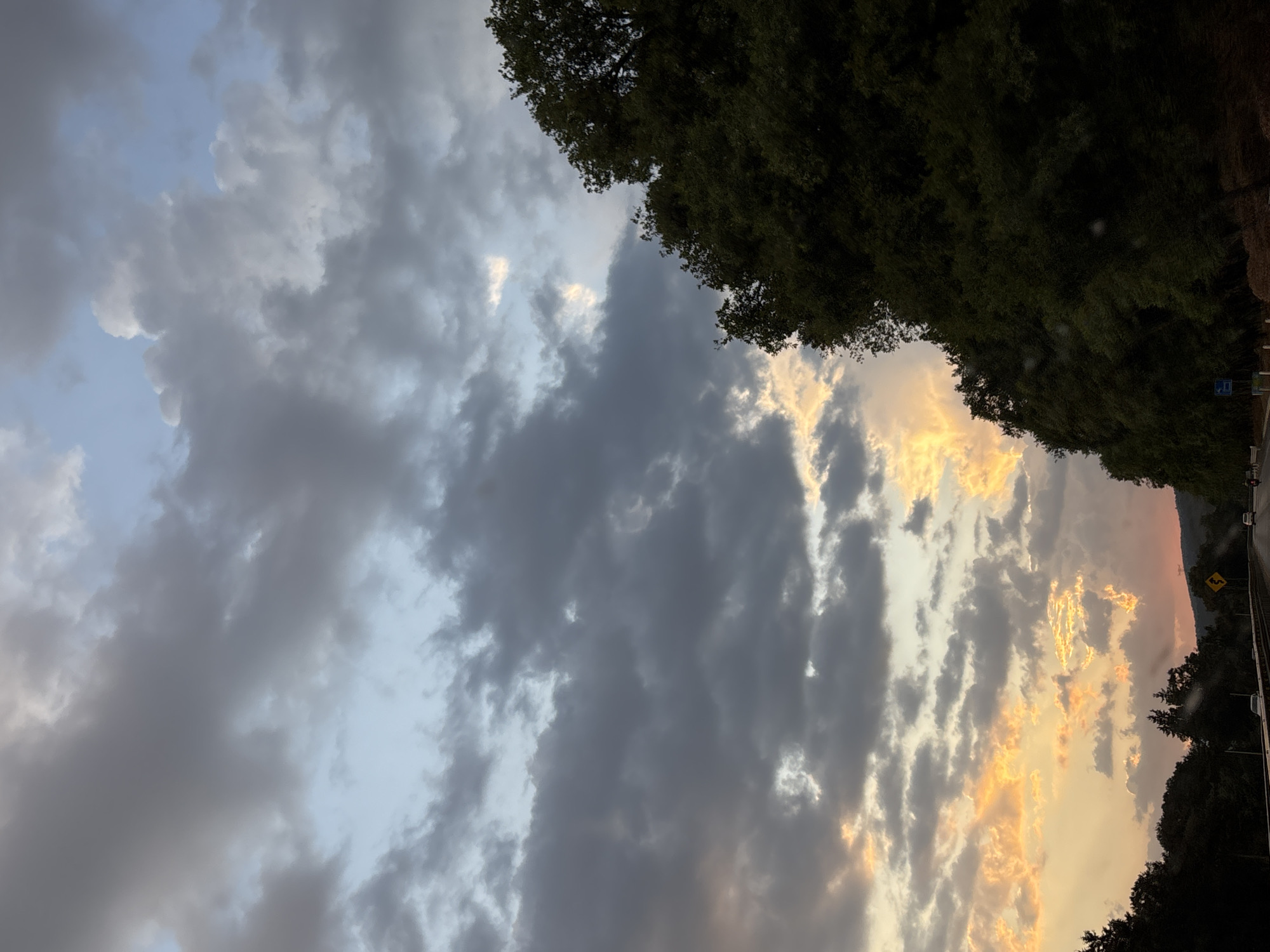 The image depicts a view of the sky with clouds, some illuminated by the setting sun, creating an orange hue. A line of trees is visible along the right edge, suggesting a landscape or roadside setting. In the bottom right corner, a glimpse of a road with a road sign can be seen. The overall scene appears to be a natural environment with some man-made elements like the road. The location is Tepoztlán, Mexico.