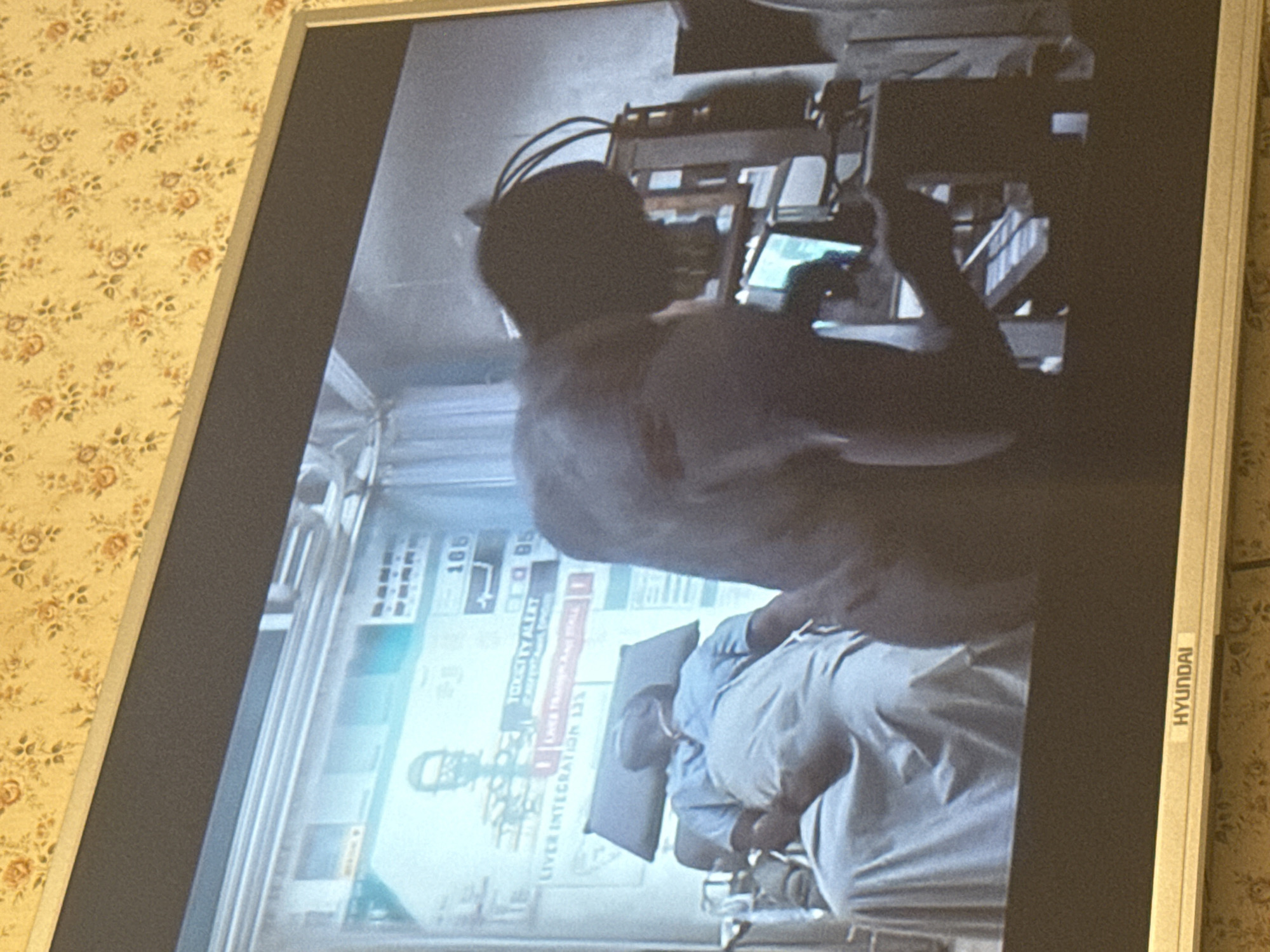 A photograph of a Hyundai television screen displays a scene featuring a man lying in what appears to be a medical or examination bed. A second individual stands over the patient, possibly attending to them or performing some kind of medical procedure. The medical environment includes monitors displaying information such as "Toxicity Alert" and "Liver Integration 133." The scene's location cannot be determined as Mytishchi, Russia from the visual information provided.