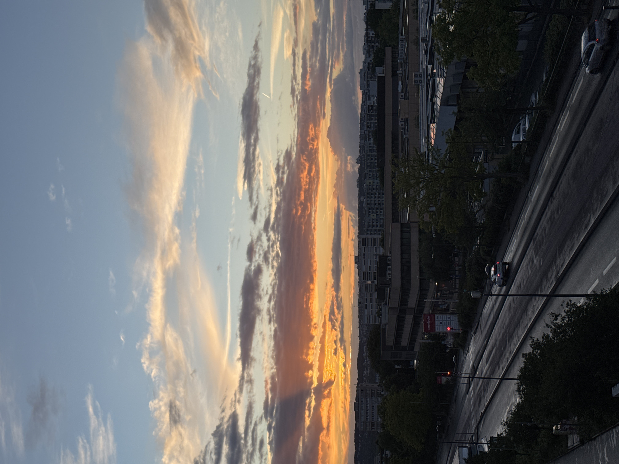 This image captures a sunset over the city of Fresnes, France. The sky is a canvas of warm oranges, pinks, and yellows, with scattered clouds tinged by the setting sun. A faint contrail from an airplane is visible high in the sky. Below the dramatic sky, the cityscape of Fresnes unfolds, characterized by mid-rise apartment buildings and commercial structures. A multi-lane road cuts through the scene, with a few cars visible, including one driving away from the viewer and another parked. Streetlights illuminate parts of the road, and trees line the sides of the street and surround some of the buildings, adding a touch of green to the urban landscape. The overall impression is of a city transitioning from day to night under a vibrant sunset.