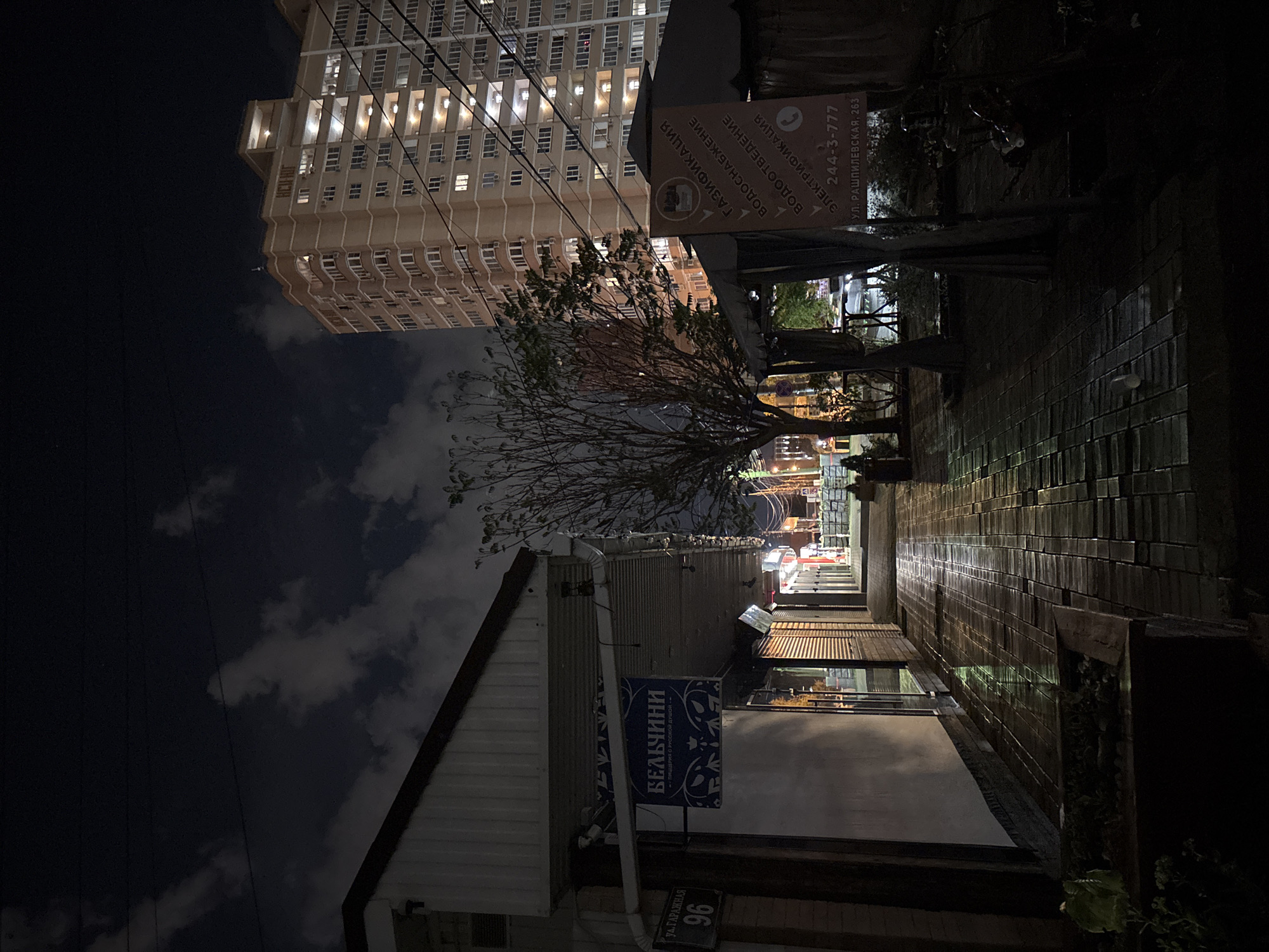 The image depicts a nighttime street scene in Krasnodar, Russia, likely in a residential or commercial area. In the foreground, a wet, paved walkway reflects the ambient light. To the left, a low-rise building with a white facade and a dark roof extends into the frame. A sign with Cyrillic text and a blue background, reading "БЕЛЧИНИ" and below it "пиццерия с русской душой", is visible on this building. A street sign indicating "ул. Гаражная 96" is also present.
Dominating the background is a tall, multi-story apartment building, illuminated from within, with numerous windows and balconies. Power lines crisscross the sky in front of the apartment building. To the right of the walkway, a covered outdoor seating area or kiosk is visible, with a sign bearing Cyrillic text and a phone number "244-3-777" along with an address "ул. Рашпилевская, 263". A bare-branched tree stands between the walkway and the kiosk. The overall impression is one of a quiet urban environment after dark, with some commercial activity indicated by the signs.