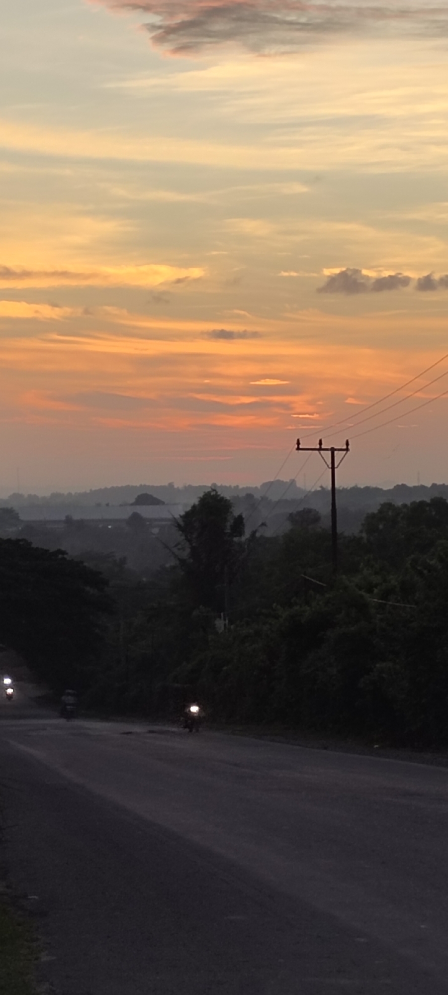 A low-angle, medium shot depicts a paved road in Lhokseumawe, Indonesia at dusk or dawn. The sky transitions from light yellow to orange hues, with a few scattered clouds.
A power pole is positioned on the right side of the frame, with wires extending across the sky. Dense foliage lines the sides of the road.
Three motorcycles are visible on the road, each with headlights illuminated. One motorcycle is closer to the camera than the others. The road surface appears dark and somewhat uneven. The overall atmosphere suggests early morning or late evening in a rural or suburban area.