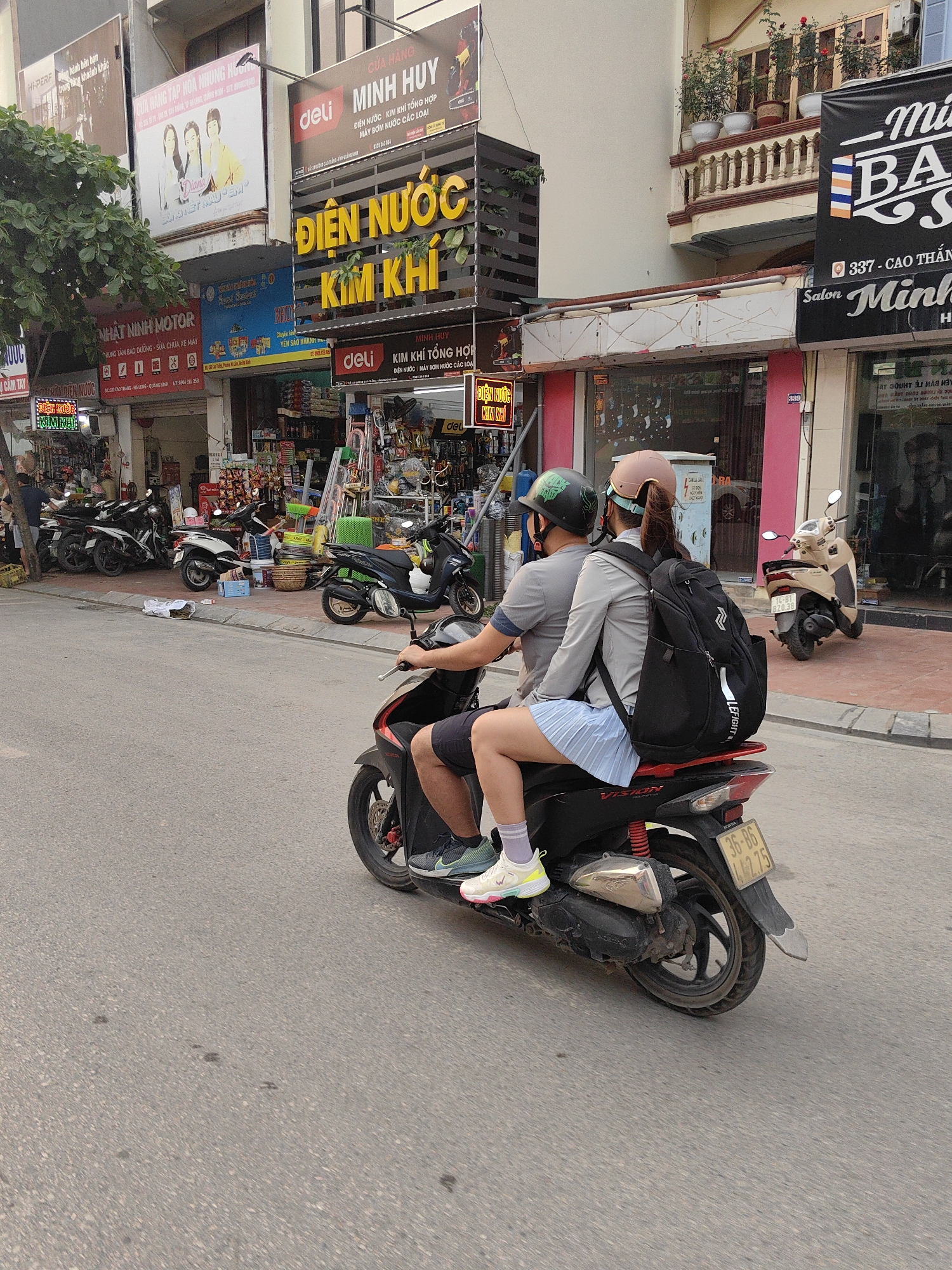 A motorcycle carrying two people is traveling on a street in Ha Lam Ward, Vietnam. The driver, a male, wears a gray shirt, shorts, and a green helmet. The passenger, a female, wears a gray jacket, a blue skirt, and a tan helmet. She is carrying a black backpack. In the background, storefronts with signs in Vietnamese are visible, including "DIEN NUOC KIM KHI" in yellow letters. Parked motorbikes line the sidewalks. The street is paved and shows signs of wear.