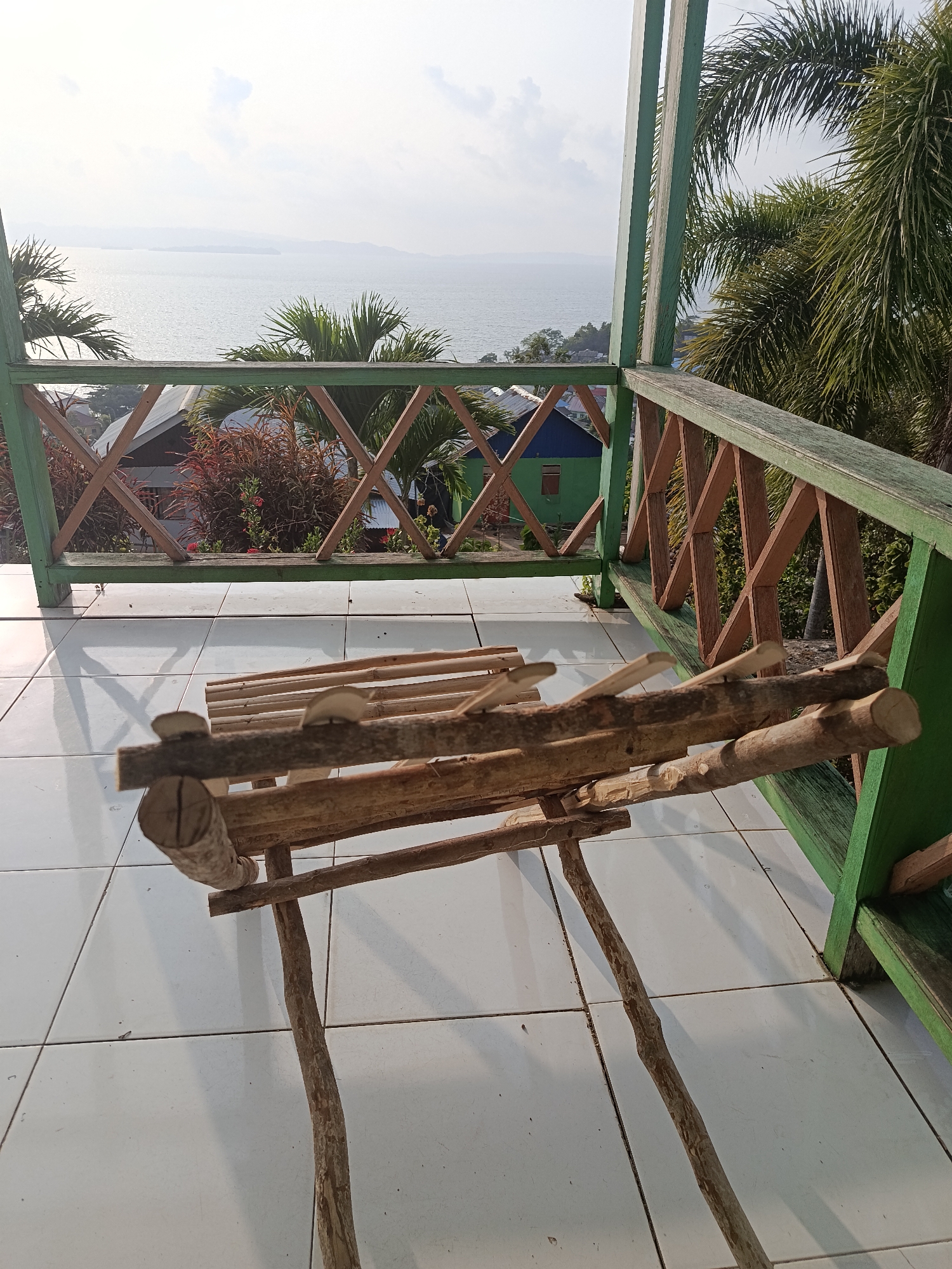 An eye-level, full shot shows a wooden lounge chair constructed from raw branches placed on a tiled patio. The chair is positioned in the foreground and is the main subject of the image. Behind the chair, a green wooden railing with a diamond pattern encloses the patio. Beyond the railing, palm trees frame the view of a calm body of water, possibly a lake or sea, under a hazy sky. In the middle ground, several buildings with red and green roofs are visible nestled among lush vegetation. The environment appears to be a serene, outdoor setting, possibly a balcony or porch overlooking a natural landscape in Mansalean, Indonesia.