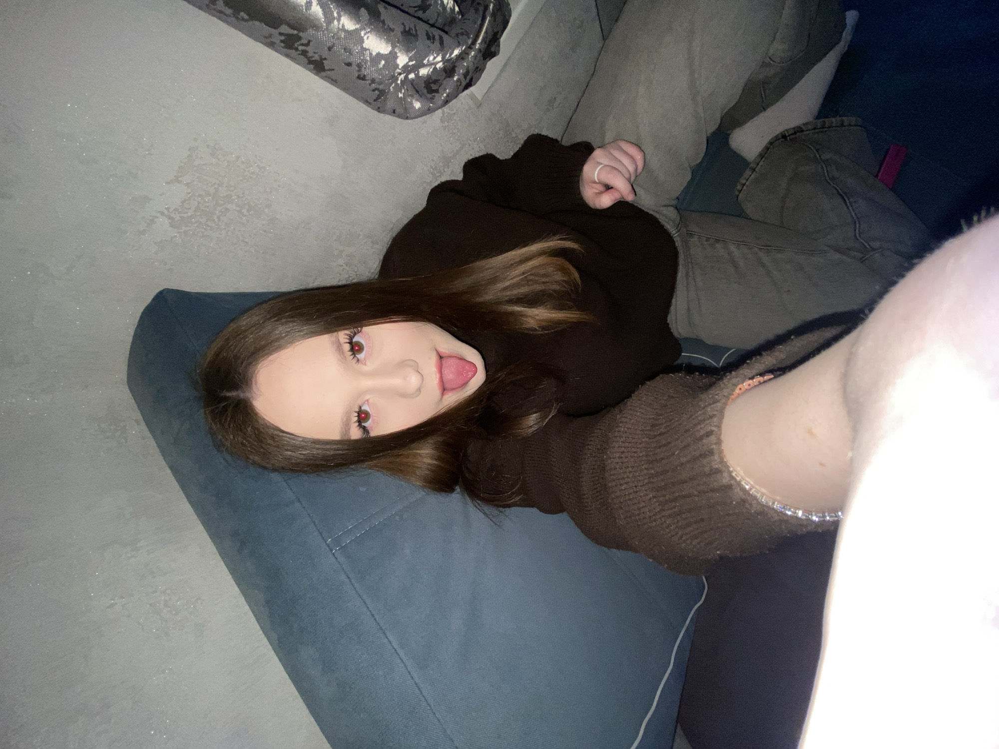 A woman is lying on a blue couch. She is wearing a dark brown sweater and grey jeans. Her tongue is sticking out. She has long brown hair. The location is not discernible from the image.