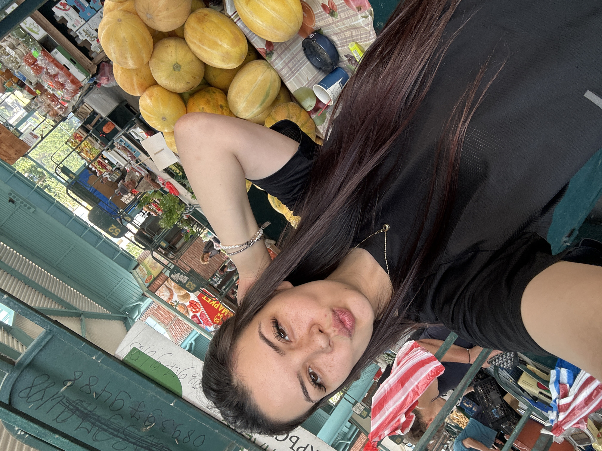 This is a selfie taken by a young woman with dark hair, freckles, and light brown eyes, who is puckering her lips. She is wearing a black t-shirt and a necklace with a small gold pendant. She has on a bracelet made of pearls and silver beads. The background is a bustling outdoor market, likely the Sliven Central Market, given the information available. Numerous stalls are visible, displaying a variety of fresh produce, including large piles of yellow melons. There are people browsing and shopping in the background, and some of the market stalls have signage in Cyrillic. The overall environment appears to be an active and vibrant marketplace.