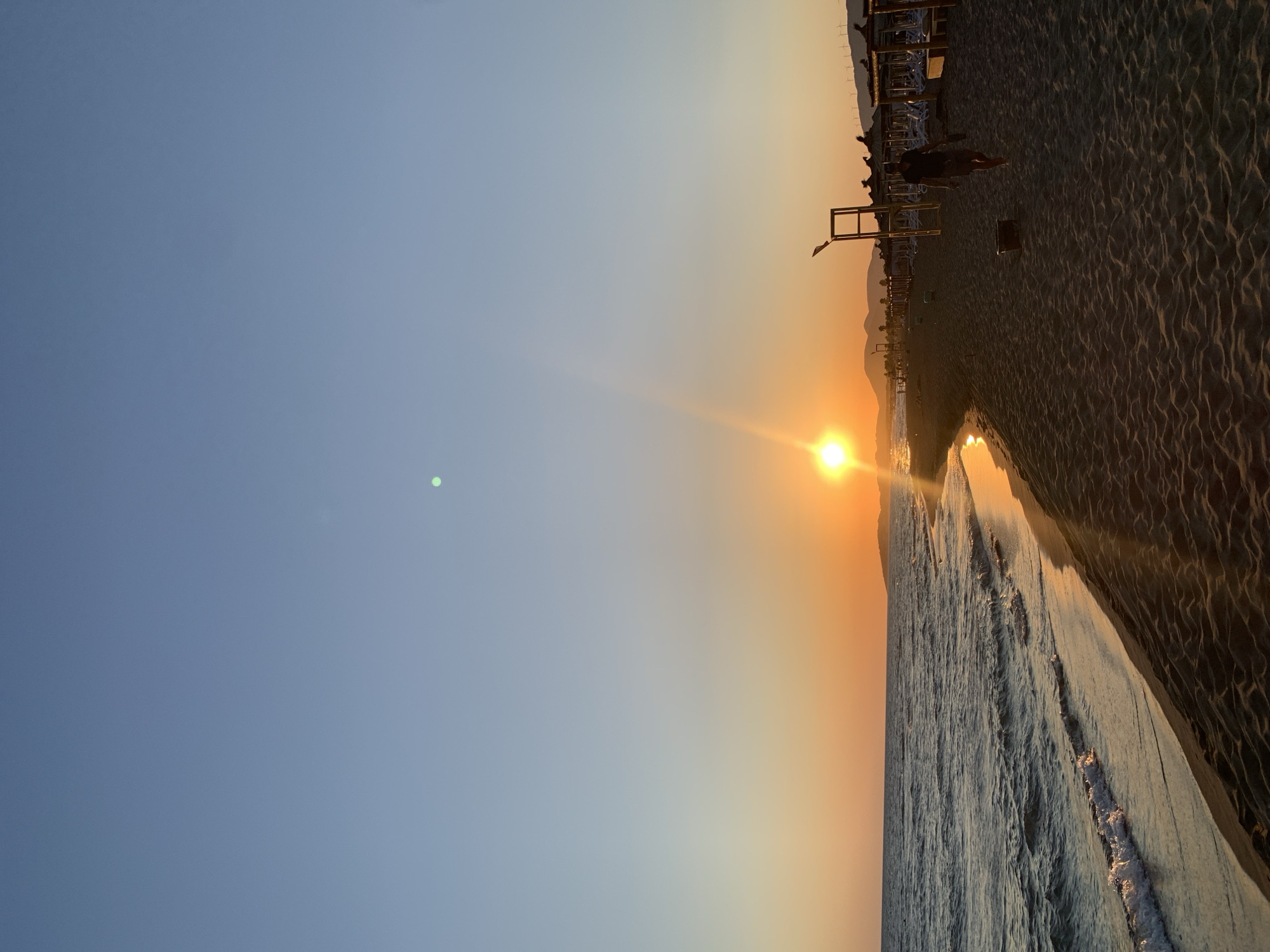 The image captures a sunset at a beach in Donji Štoj - Shtoji i Poshtëm, Montenegro. The sun is low on the horizon, casting a warm orange glow across the sky and water. Gentle waves lap at the sandy shore, which is lined with beach umbrellas and loungers. A solitary figure walks along the beach, heading away from the viewer, towards the setting sun. The scene is serene, with the vast expanse of the sea meeting the clear sky, creating a picturesque end to the day.