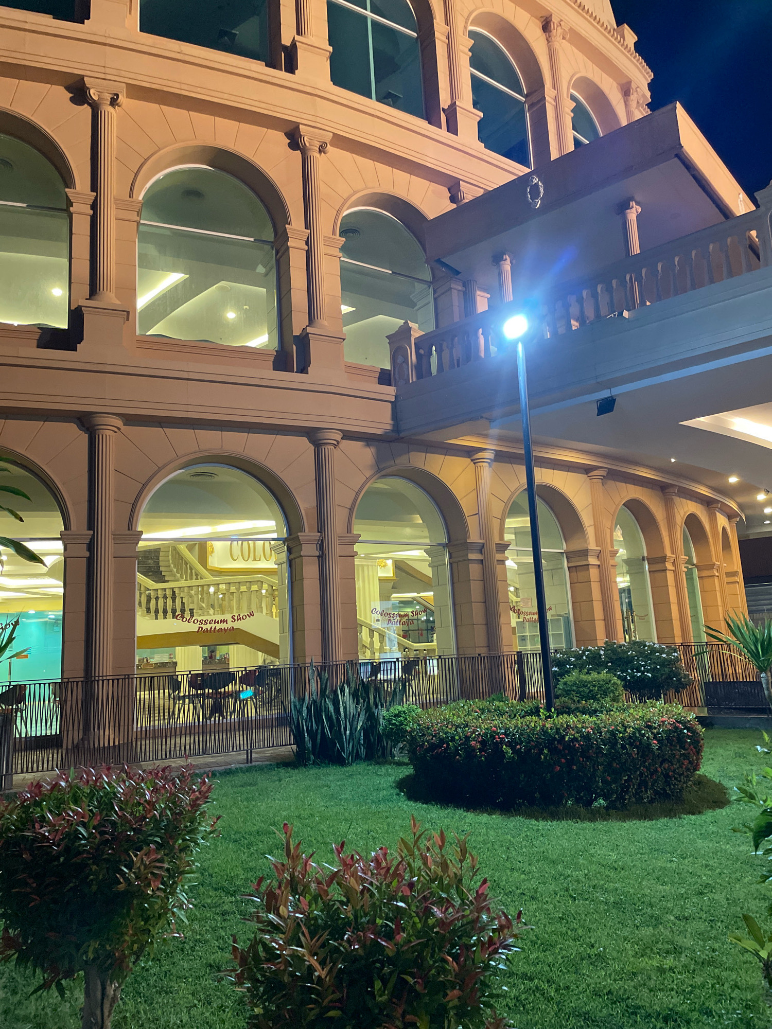 The image shows the exterior of the Colosseum Show Pattaya in Pattaya City, Thailand, at night. The building features classical architecture with arches, columns, and a prominent balcony. The facade is illuminated, highlighting the architectural details and the surrounding landscaped garden with manicured hedges and grass. A street lamp is visible in the foreground, casting a bright light. Through the arched windows, a grand staircase and signage for "Colosseum Show Pattaya" are visible, suggesting an entertainment venue. There are no people or active events depicted in the scene. The overall impression is that of an impressive and well-maintained entertainment complex at rest.