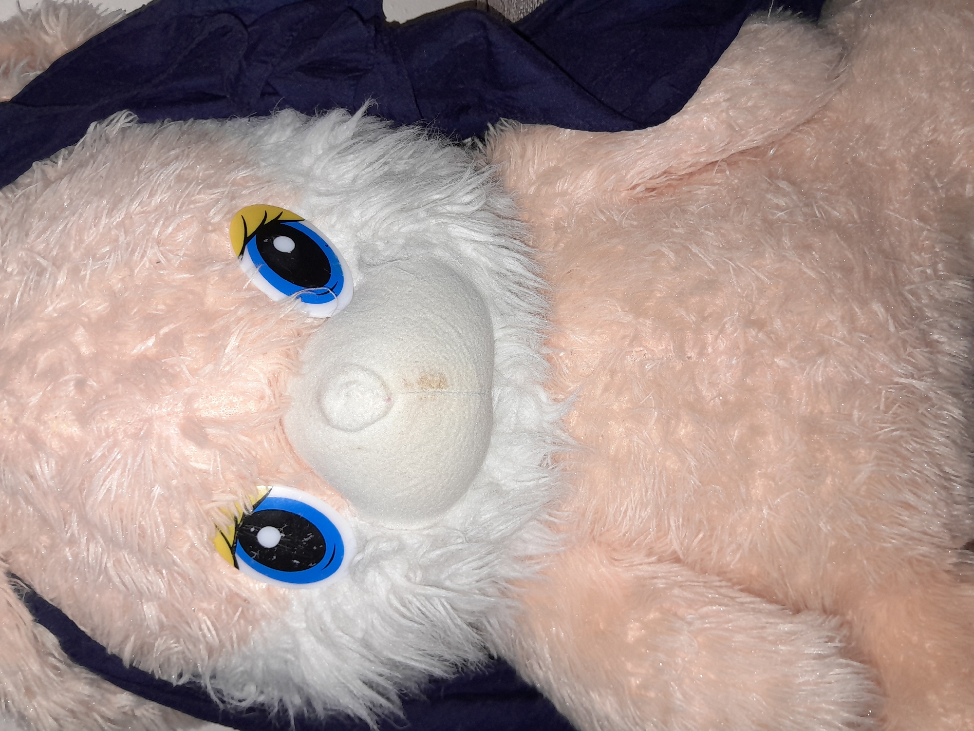 The image is a close-up, high-angle shot of a stuffed animal, likely a bear or rabbit, with predominantly pink and white fur. The stuffed animal has large, round blue eyes with black pupils and white highlights, outlined in black. Above each eye, there is a yellow shape representing an eyelid or eyebrow. The nose is a small, white, rounded bump. A faint brown stain is visible on the stuffed animal's muzzle. A dark blue fabric is draped behind and partially to the side of the stuffed animal. The lighting appears to be artificial, casting some reflections on the fur, which has a slightly glittery appearance. The background is out of focus, suggesting the stuffed animal is the primary subject. There are no people or discernible environmental features related to the city of Uchquduq, Uzbekistan, present in the image.