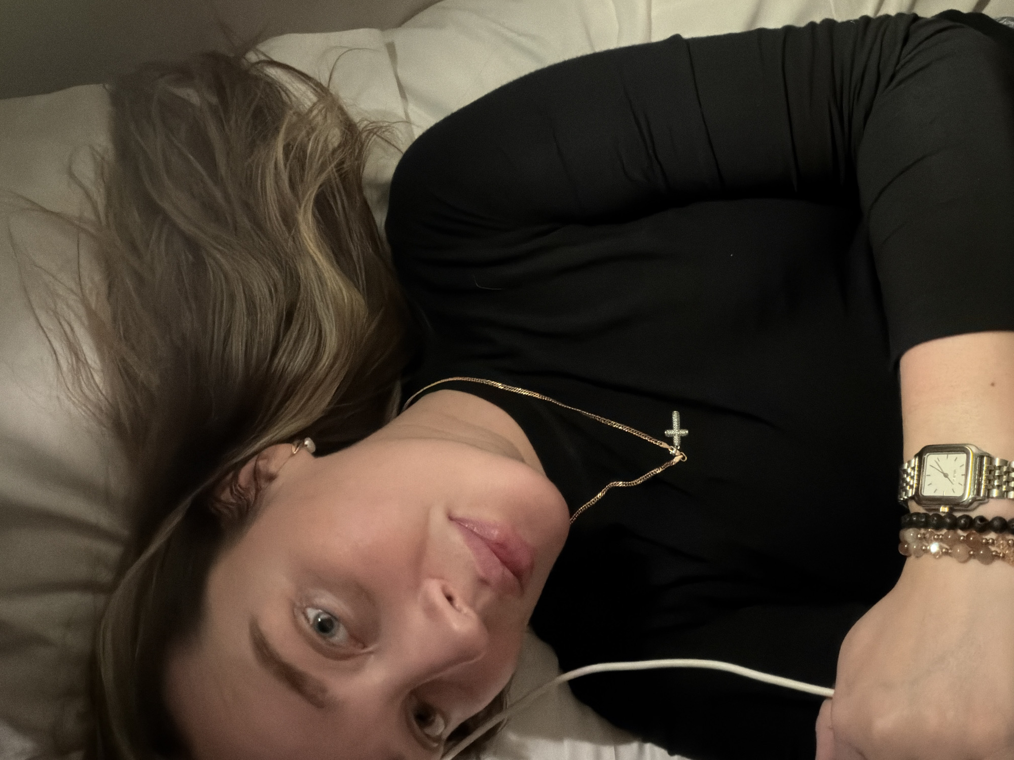 A woman is lying on her back on a white pillow, likely in a bed. She is wearing a black long-sleeved shirt, a gold necklace with a cross pendant, a wristwatch, and bracelets. Her hair is light brown with blonde highlights. Her gaze is directed towards the camera. The environment appears to be indoors, possibly a bedroom. There is no indication of the city of Moscow or any other specific location.