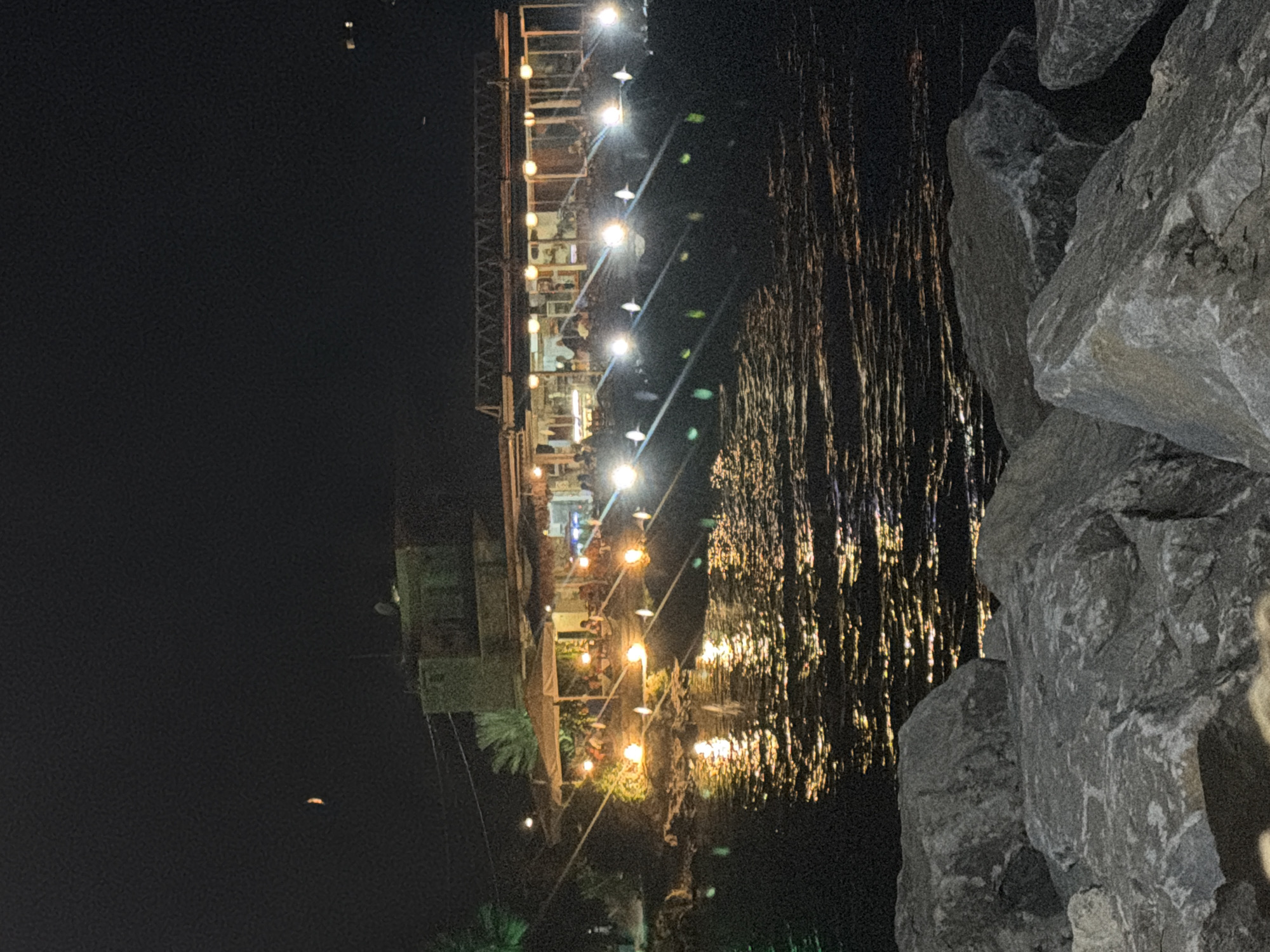 The image shows a waterfront restaurant in Georgioupoli, Greece at night. The restaurant is lit with string lights and has a covered outdoor seating area where patrons are visible. The water in front of the restaurant reflects the lights, creating a shimmering effect. Large rocks are in the foreground, obscuring the bottom of the image. A sliver of the moon is visible in the dark sky above.