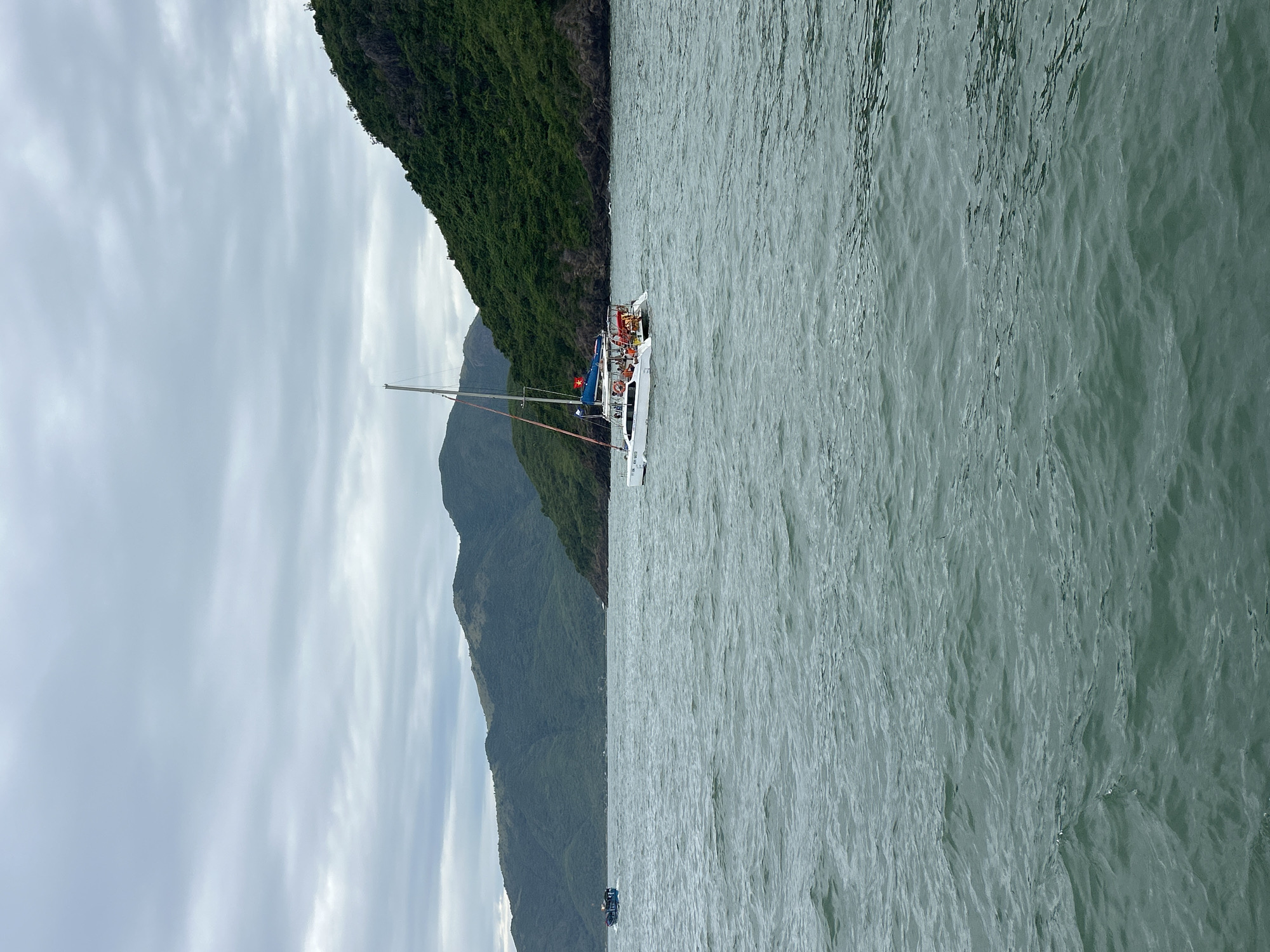 A white catamaran, carrying several passengers wearing life vests, sails on choppy, dark green water. In the background, lush green hills form a backdrop to the ocean. Another smaller, dark-colored boat is visible in the distance to the left. The sky is overcast with gray clouds. This scene likely depicts a boat tour or a leisure activity in the waters off the coast of Nha Trang, Vietnam.