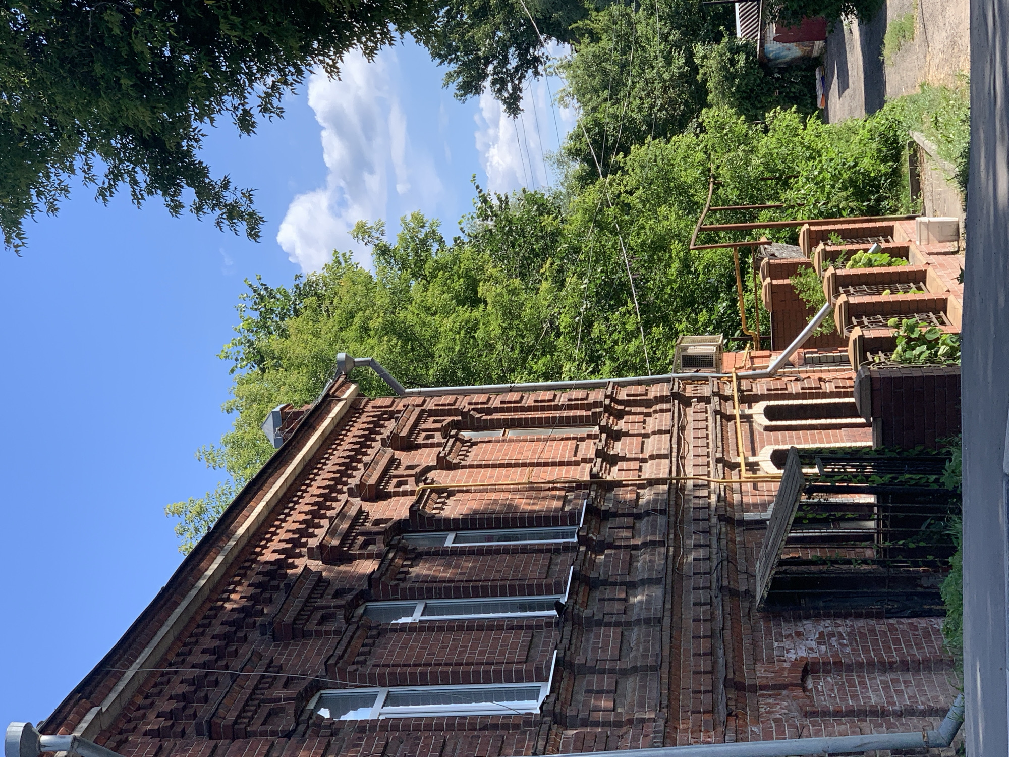The image depicts an old brick building with ornate architectural details, including decorative brickwork along the roofline and around the windows. Several tall, narrow windows with white frames are visible on the building's facade. A metal downspout runs along the side of the building. In front of the building, there is a fenced-off area with a small, angled wooden roof. Green vines are growing on the fence. A yellow pipe runs vertically up the side of the building, and a small air conditioning unit is mounted on the wall.
To the right of the building, a pathway or driveway leads away, flanked by lush green trees. The sky is a clear blue with scattered white clouds. The overall impression is of an older, possibly historic, residential building in a semi-urban or suburban setting, with abundant greenery. The image appears to be taken on a sunny day.