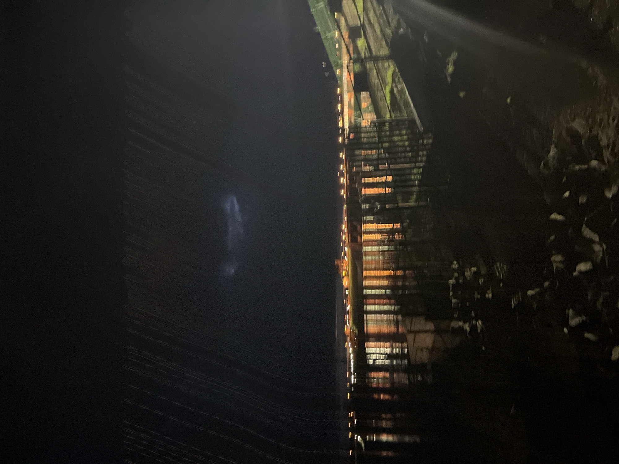 The image presents a nighttime scene overlooking the Votkinsk Reservoir of the Kama River in Chaikovsky, Russia. The sky is dark, with numerous diagonal streaks in the upper left quadrant consistent with falling rain or light artifacts from precipitation. A faint, diffuse lighter area, possibly an illuminated cloud, is visible in the upper center sky.
Across the reservoir, a distant shoreline is visible, characterized by a concentration of artificial lights, primarily orange and white, which are blurred and reflected in elongated patterns on the water's surface. These lights suggest the presence of urban or industrial infrastructure.
In the midground, a dark, vertical grid-like structure, resembling a fence or a series of support pilings, extends into the water, casting dark reflections. The immediate foreground on the left depicts dark, uneven water or wet ground containing scattered light-colored debris. On the right side of the foreground, a dark, sloped embankment with sparse vegetation is partially visible, illuminated by a localized, intense light source that creates a lens flare in the lower right corner of the frame. No individuals are present in the image.