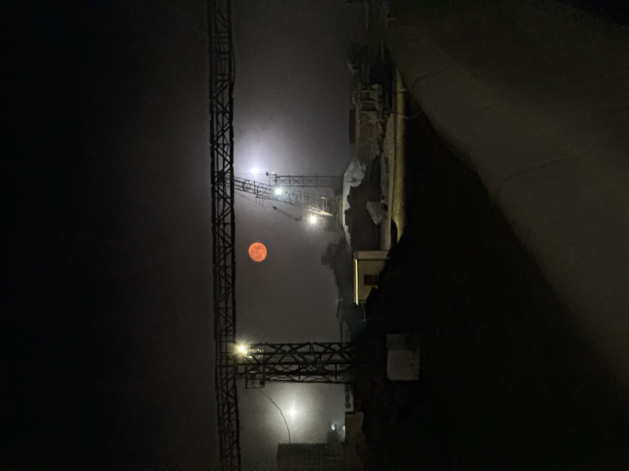 The image depicts a construction site at night under a hazy, possibly foggy, sky. A large, reddish-orange moon is visible in the upper center of the frame, partially obscured by the mist. Several construction cranes and industrial structures are present. A prominent horizontal truss structure spans across the upper portion of the image. On the left, a tall, dark-colored metal tower with lights is visible, possibly part of a crane. Further to the right, another crane structure rises from the mist. Piles of dirt or debris are scattered throughout the foreground and midground. A small, illuminated structure with a doorway is situated to the right of the central tower. The overall atmosphere is dark and industrial, with artificial lights from the site cutting through the atmospheric haze and casting an ethereal glow. The scene is devoid of people or any discernible activity. The location is identified as Magnitogorsk, Russia.