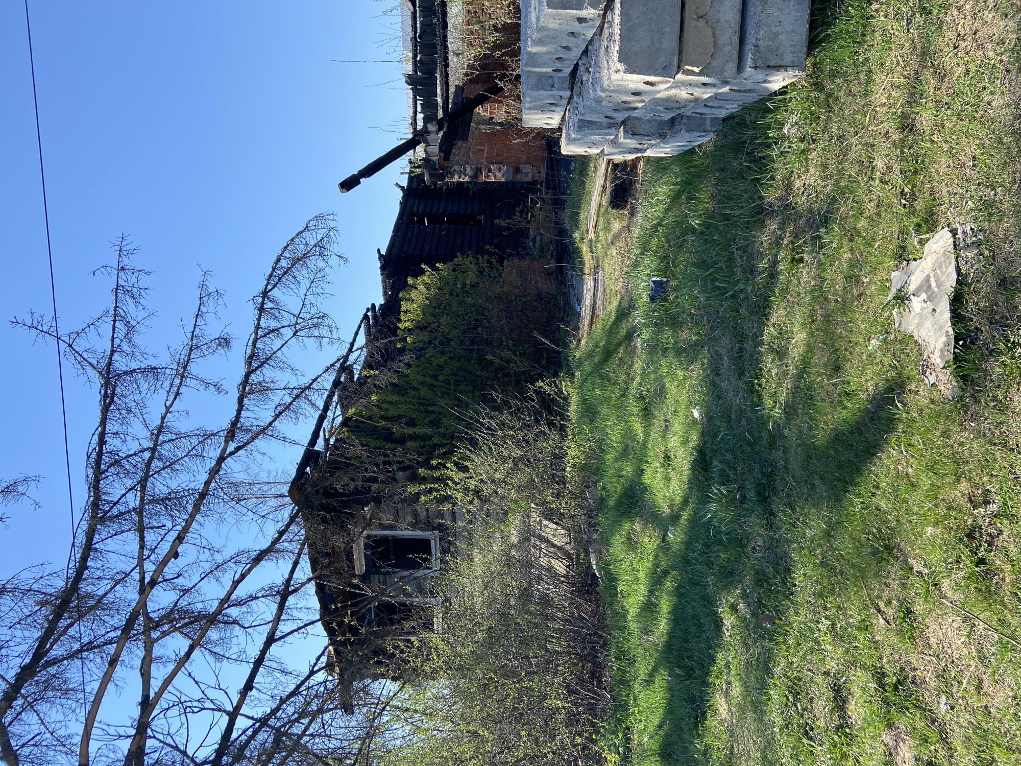 The image depicts the aftermath of a fire in what appears to be a residential structure. The building is severely damaged, with charred remains of wood and brick visible. A portion of the roof has collapsed, and the window frames are blackened. The surrounding vegetation is green, suggesting the incident occurred during a period of growth. Concrete blocks are stacked near the building. The sky is clear blue. The location is Chaikovsky, Russia.