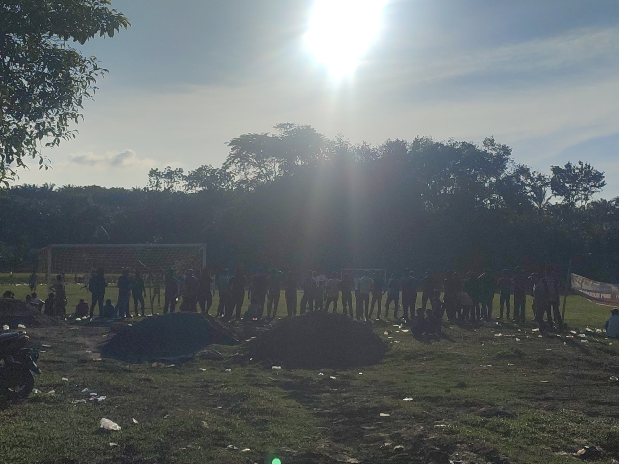 The image shows a group of approximately 30 people gathered on a grassy field in Babahlueng, Indonesia. A soccer goal is visible in the background, along with trees and a bright sun. In the foreground, there are piles of dark material, possibly soil or gravel, and scattered trash. A motorcycle is parked on the left side of the frame. The people appear to be standing or sitting.