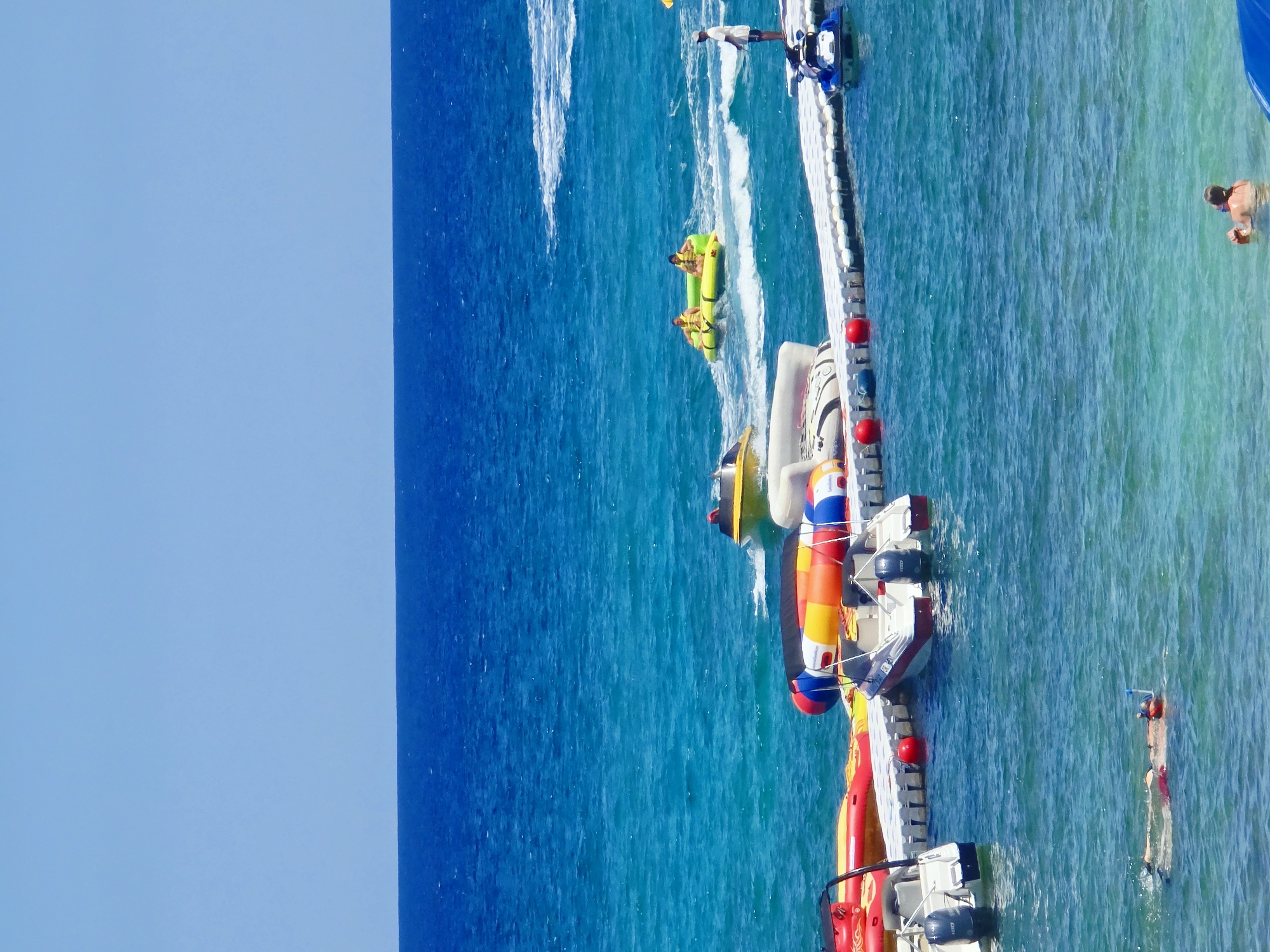 The image captures a lively scene at the coast of Protaras, Cyprus, on a bright, sunny day. The turquoise waters of the Mediterranean Sea are dotted with various watercraft and recreational activities. In the foreground, a person is snorkeling, with their head submerged and snorkel visible. A bit further out, another person is partially visible in the water, holding onto a floating object.
A white modular dock stretches out into the water, serving as a platform for several inflatable water toys and two small motorboats. One motorboat, white with red accents, is docked at the end of the platform. Another motorboat, primarily yellow, is seen further in the distance. On the dock itself, there are colorful inflatable tubes.
In the mid-ground, two people in life vests are riding on a yellow inflatable tube being pulled by an unseen boat, creating a wake behind them. A man in a white shirt and shorts stands on the far end of the dock, observing the activities. A blue and white jet ski is also moored to the dock. In the background, the vast expanse of the deep blue sea meets the clear, pale blue sky, with a faint white wake visible on the horizon. The overall impression is one of a recreational resort area, bustling with holiday activities under a clear sky.