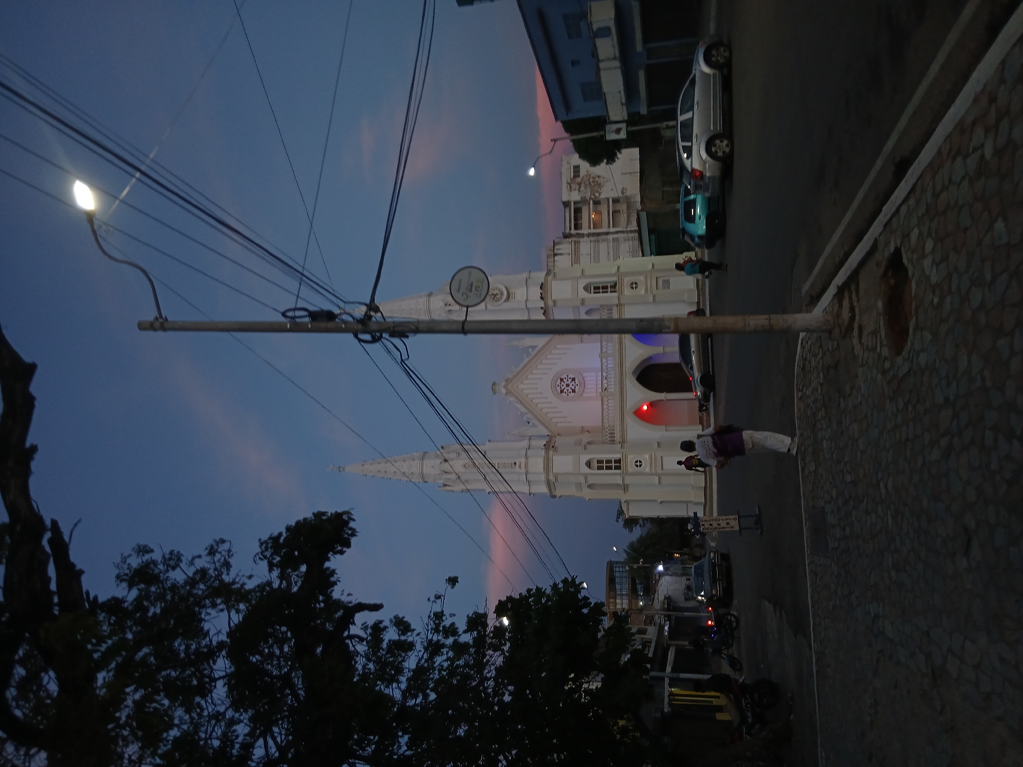 The image captures an evening street scene in Juan Griego, Venezuela, centered around a prominent white church under a twilight sky. The sky displays a gradient of deep blue transitioning to lighter shades with hints of pink and orange clouds, indicating either dusk or early evening. A bright streetlight is visible on the left, casting light onto the overhead power lines that crisscross the upper part of the frame.
The main subject is a large, ornate white church, likely a cathedral, with a tall steeple, multiple arched windows, and a distinctive rose window above its main entrance. The church's facade appears illuminated, highlighting its intricate architectural details. On the right side of the image, a stone-paved sidewalk or wall runs alongside a road where several vehicles are present, including a silver car, a light blue car, and a motorbike further down the street.
Several people are visible, though not clearly identifiable. One person wearing a purple top and white pants stands near the illuminated church entrance, while another in a blue top is near a parked car. Dark silhouettes of trees occupy the lower left and center-left of the frame, partially obscuring the sky. A utility pole with numerous cables stands prominently in the mid-ground, and a circular sign, possibly a pedestrian or traffic sign, is attached to it near the church.
The activity depicted is that of a calm evening in a town, with people walking and vehicles on the street. There are no overt events taking place, but rather the ambient atmosphere of daily life transitioning into night. The weather appears clear and serene. No distinct text is legible on any of the visible signs.