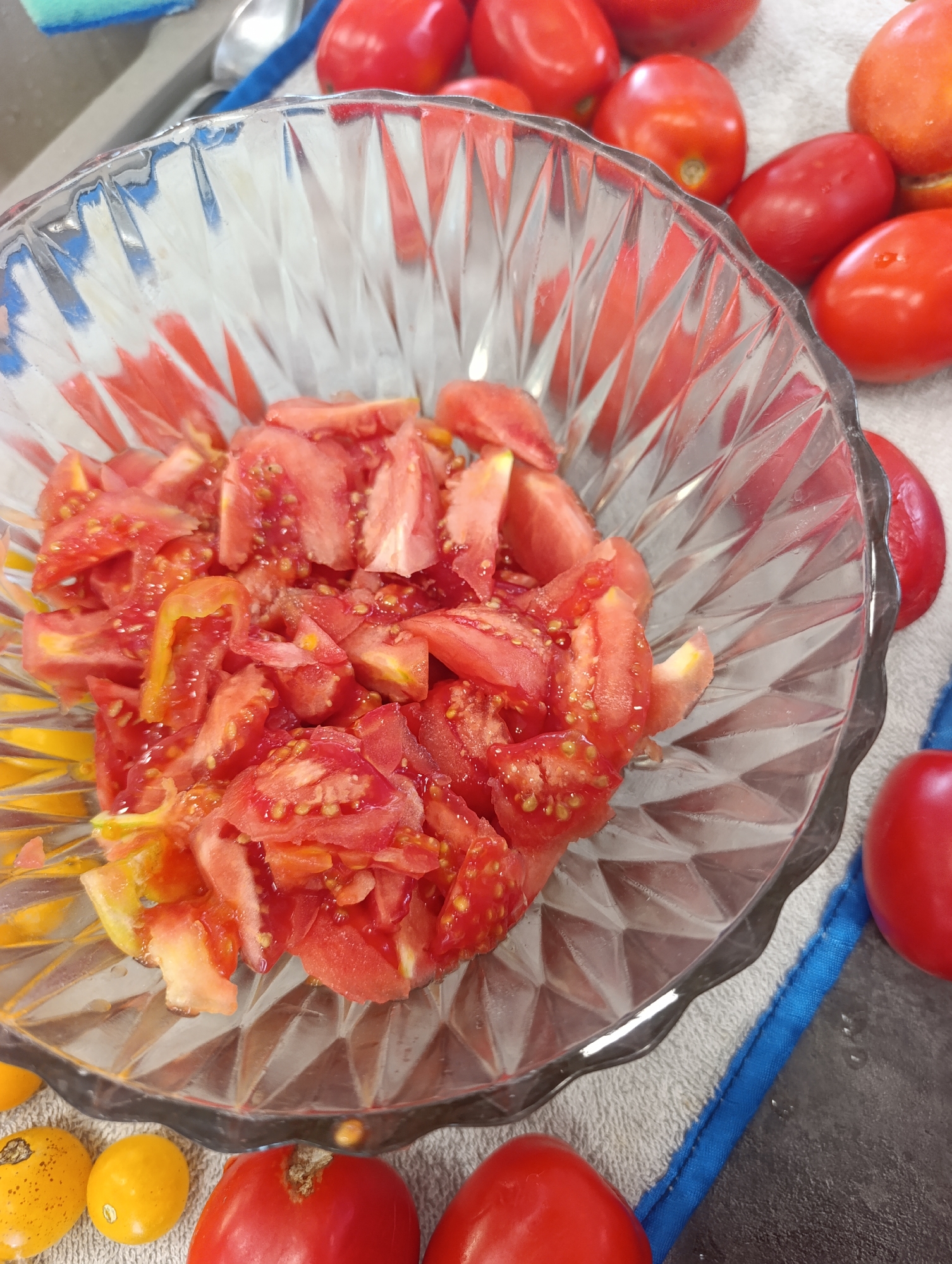 This is a close-up, high-angle, eye-level shot of chopped tomatoes in a clear, faceted glass bowl. The bowl is placed on a white textured towel with a blue edge. Several whole red tomatoes are scattered around the bowl. In the lower left, two small yellow tomatoes are visible. In the background, a blurry blue sponge and a metal spoon can be seen. The scene appears to be in a kitchen. There is no indication of the city of Ivanovka, Russia.