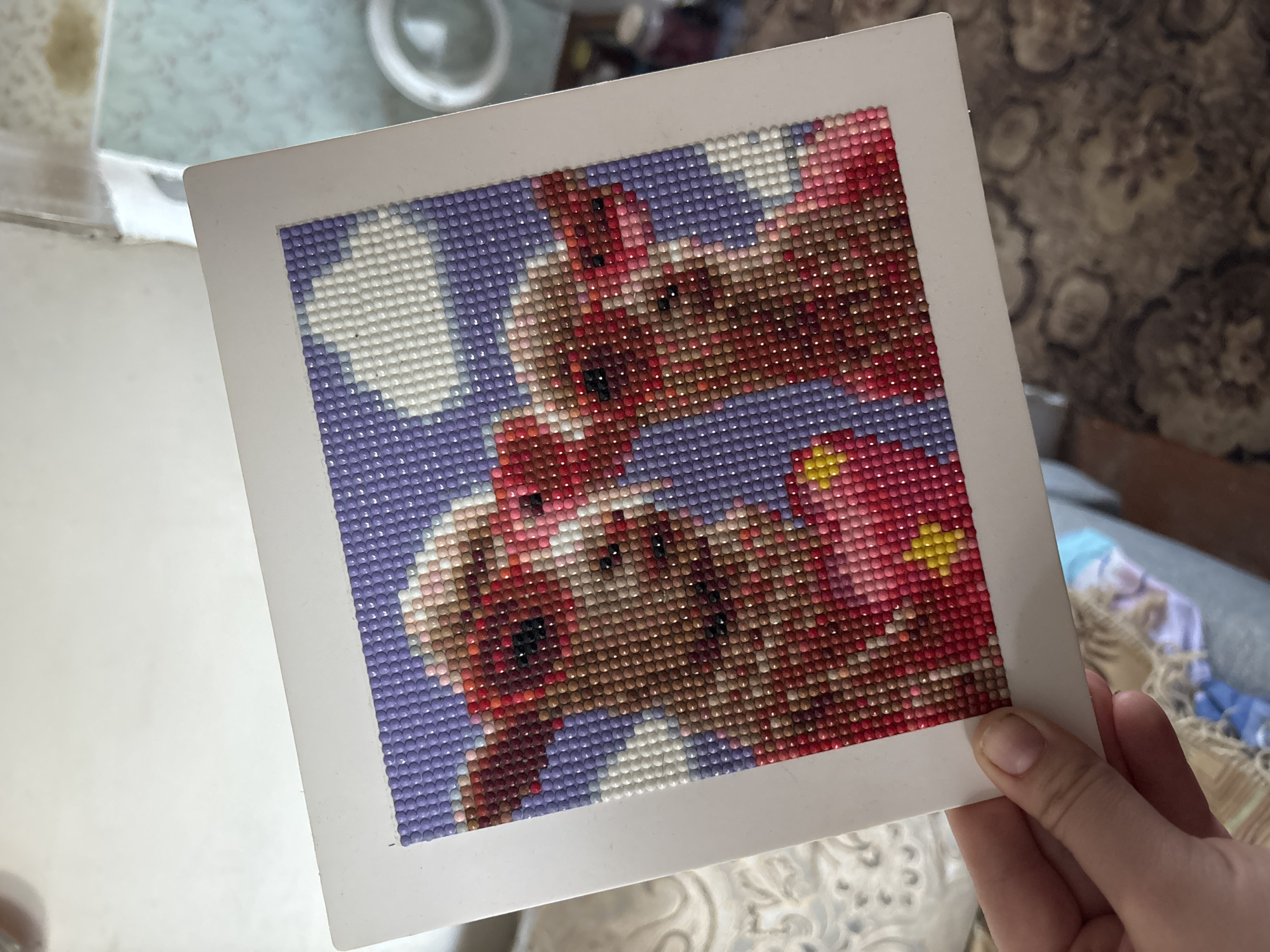 The image shows a completed diamond painting kit held in a person's hand. The painting depicts a pair of teddy bears against a background of purple sky and white clouds. The teddy bears are rendered in shades of brown, red, and pink. The painting is mounted on a white frame. The background of the photograph includes a white surface and glimpses of a patterned carpet or upholstery. The location is presumed to be Niva, Russia, but there is no specific detail confirming this.