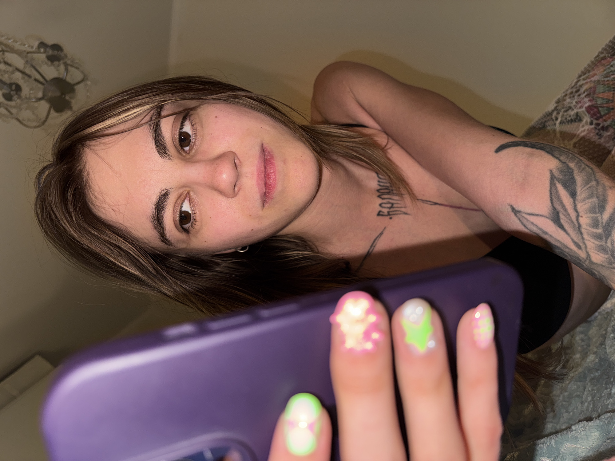 The media file is a close-up, low-angle photograph of a young woman holding a purple smartphone. She is lying down, with her head angled towards the camera, and her body is partially visible, suggesting she is in a relaxed or intimate setting. The woman has brown hair with blonde highlights and is wearing a black top. She has tattoos on her arm and chest, and her fingernails are painted with colorful designs, including green, white, and pink. The background appears to be a bedroom, with a patterned blanket visible and a chandelier or light fixture in the upper left corner. The lighting suggests it is nighttime or indoors with artificial light. The woman's expression is neutral, and she appears to be looking at the phone or the camera. There is no visible text in the media file. The overall impression is that of a casual, personal moment captured by the woman herself.
