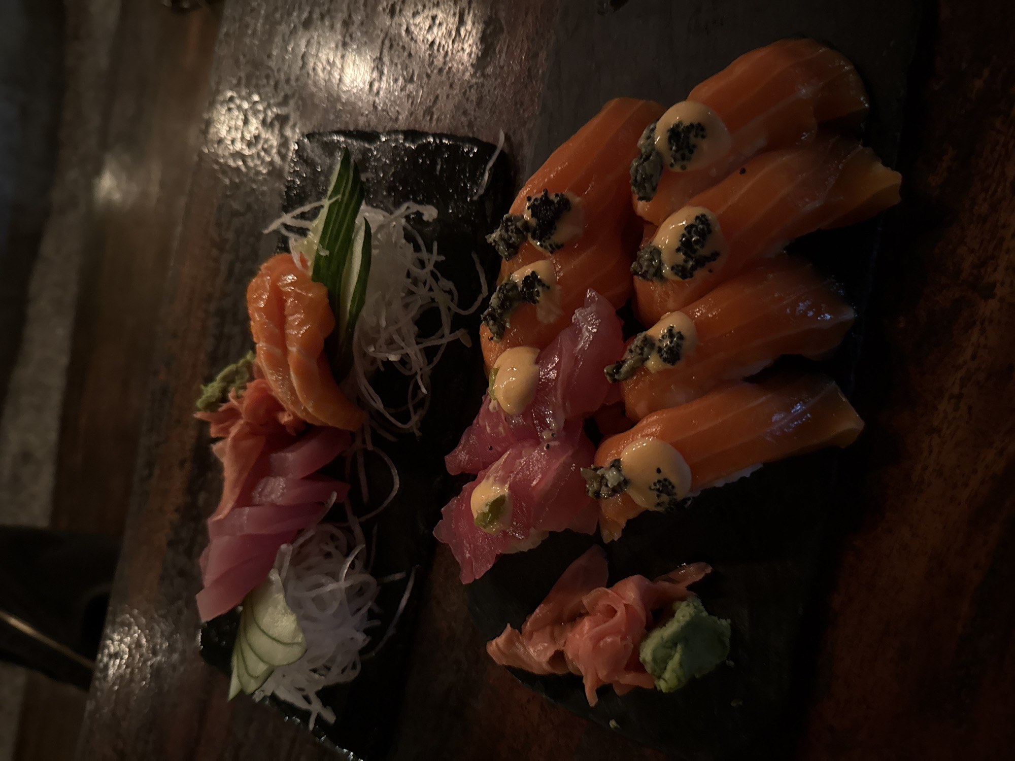 The image depicts two black plates of sushi, likely in a restaurant or food establishment. The plates are placed on a dark, possibly wooden surface.
On the plate to the left are slices of salmon and tuna sashimi, garnished with pickled ginger, wasabi, sliced cucumber and shredded daikon radish.
The plate on the right contains topped salmon and tuna sushi, also with pickled ginger and wasabi. The sushi is topped with a light-colored sauce and black sesame seeds.
Based on the prompt information, this image was likely taken in Kuta Selatan, Indonesia.