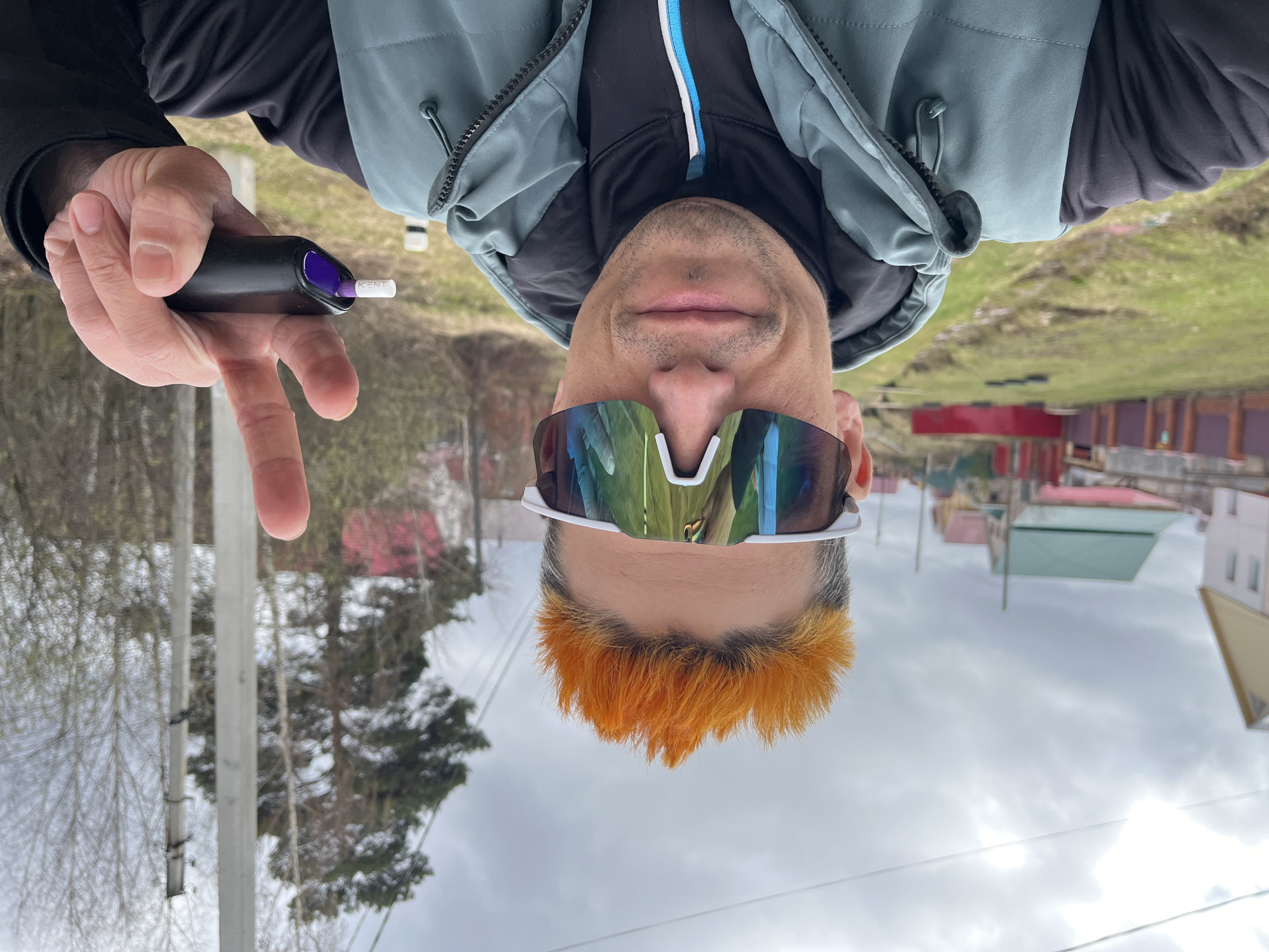 A selfie of a man in a grey zip-up vest and white athletic sunglasses, oriented so his head points downward. He has a black electronic device with a purple-tipped white cigarette in his hand. The man has orange-dyed hair. The background suggests an urban or suburban environment with buildings and trees, possibly in Chaikovsky, Russia.