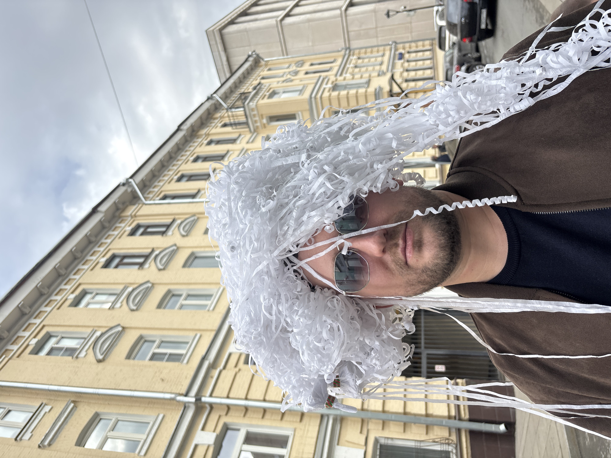A man is centered in the frame, wearing a large, white, shredded paper wig and round-rimmed sunglasses. He is also wearing a brown jacket over a dark blue shirt. The background is a multi-story, yellow building with white trim. The building is characteristic of architecture found in Moscow, Russia. The angle suggests the photo was taken from a low perspective, tilting upwards. In the upper right corner, there is a glimpse of a black car. The sky is overcast.