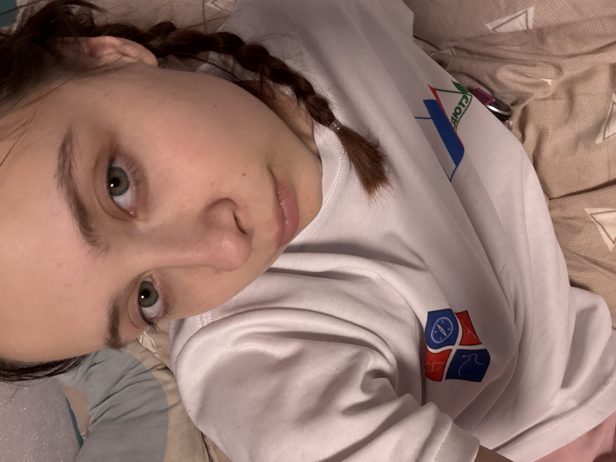 A young, light-skinned woman with braided brown hair lies on a bed with tan patterned sheets. She is wearing a white t-shirt with a logo depicting the flag of Прикамский, Russia. The logo has a blue rectangle with the letters "ДЮОТЗ" and a green branch above it. Another logo on the bottom right of the shirt has a blue compass, red mountain and blue wave.
